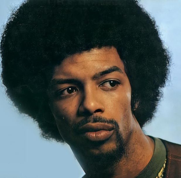 Close-up portrait of a man with dark skin, an Afro hairstyle, and a mustache and goatee, looking to the side with a serious expression, against a plain blue background.