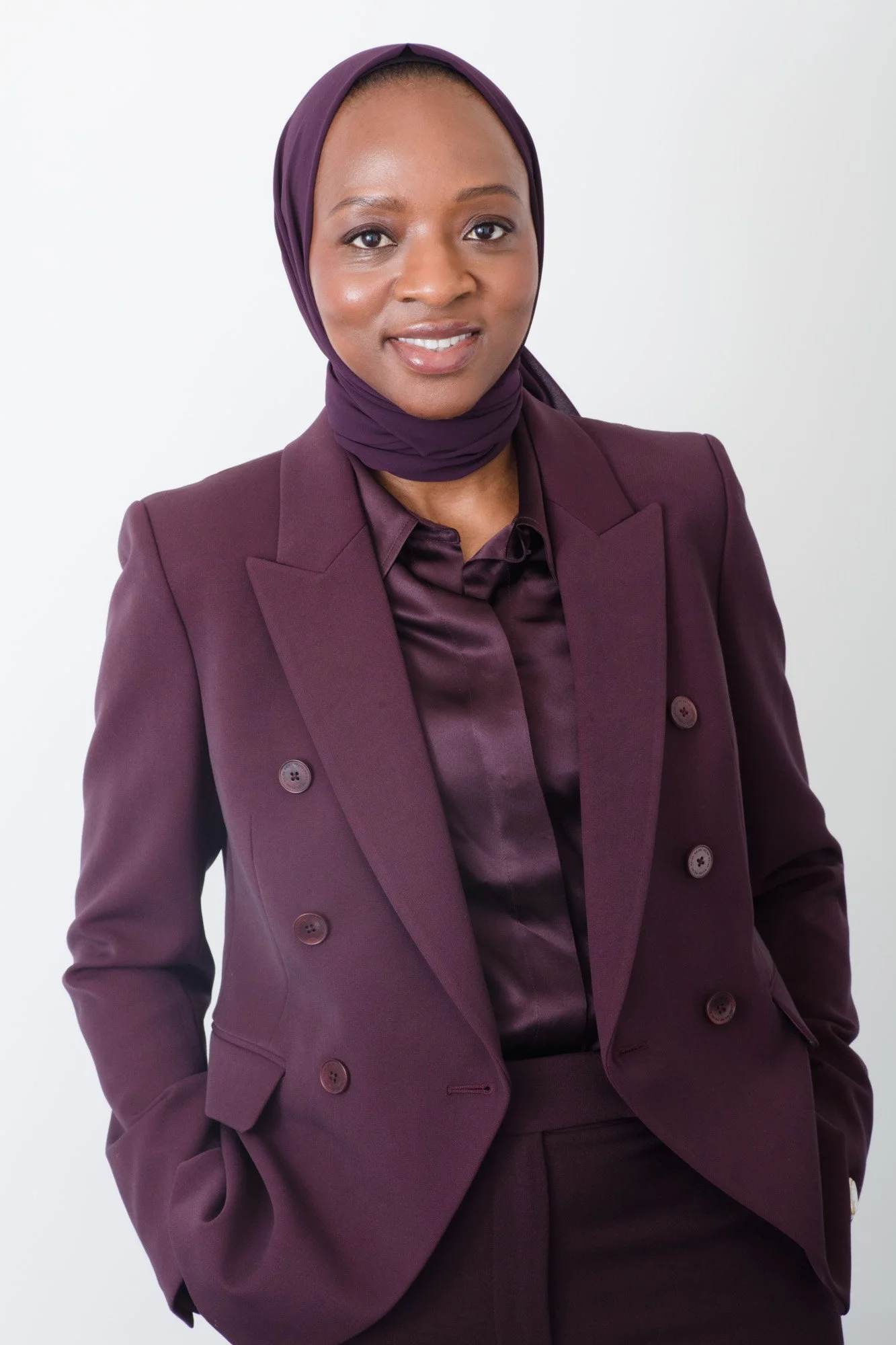 A woman wearing a purple suit with a matching purple headscarf, standing against a plain white background.