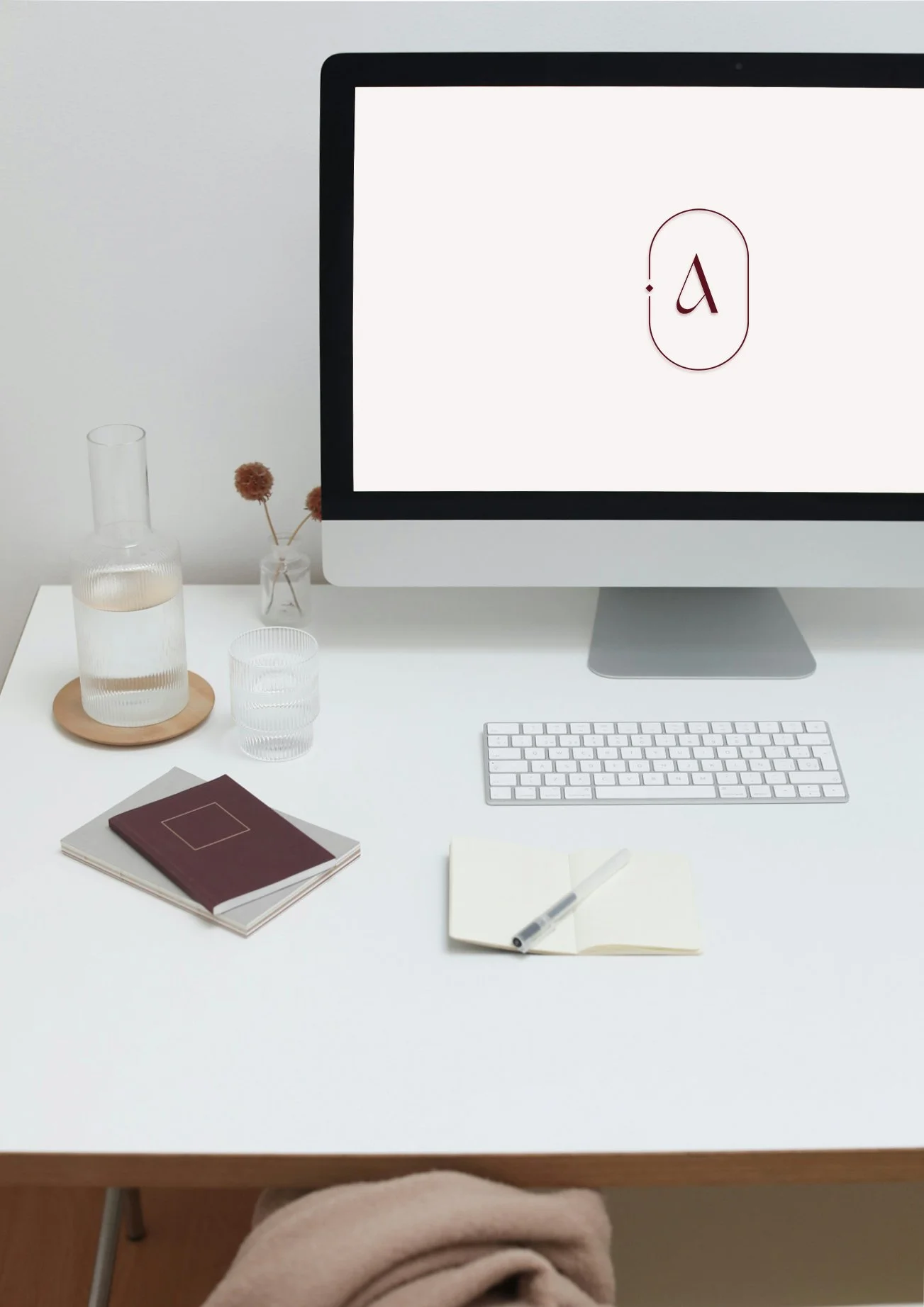 A white desk with a computer monitor displaying a simple logo, a keyboard, and a mouse. On the desk, there are two notebooks, an open notebook with a pen, a stack of books, a glass carafe and a glass of water, and a small vase with dried flowers.