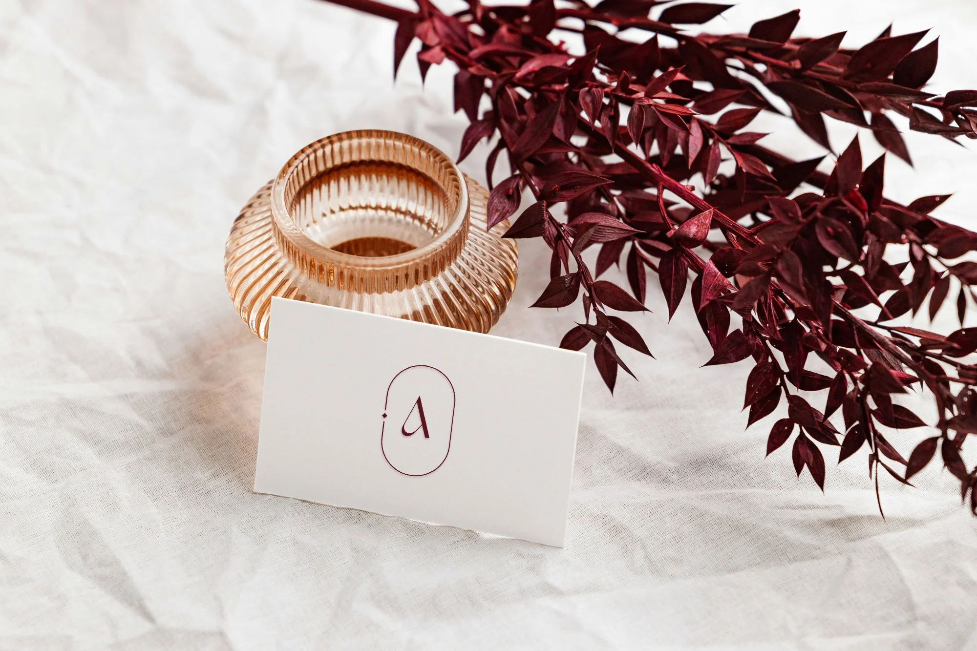 A glass vase with ribbed design and a warm hue, a burgundy-colored leafy branch, a white tablecloth, and a white card with a red number '9' printed on it.