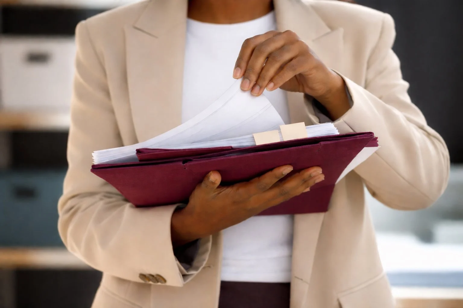 A person in a beige blazer and white shirt is holding a red binder filled with papers in an office setting.