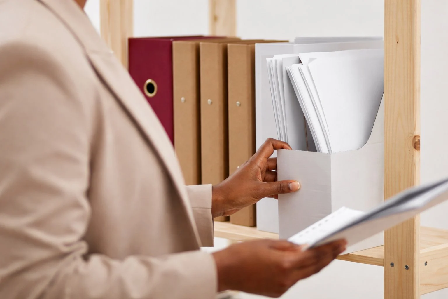 A person in a beige jacket holding a white file organizer with papers, with shelves containing red, brown, and white binders in the background.