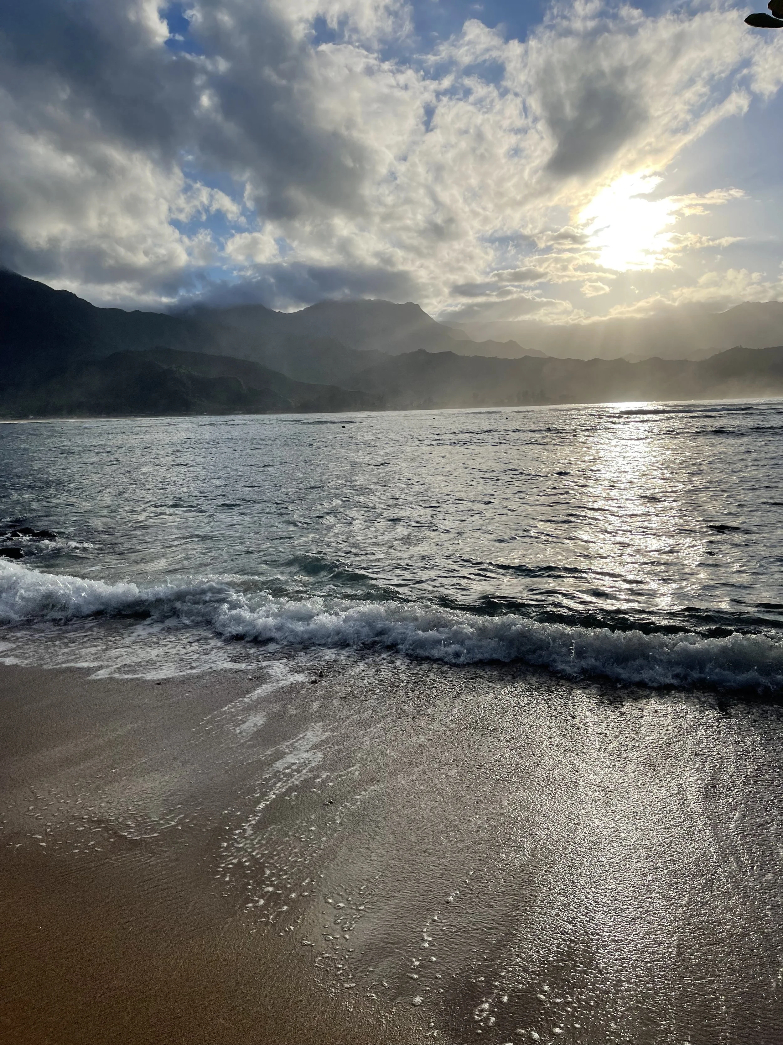 A scenic ocean view with the sun partially hidden by clouds, mountains in the background, and waves gently crashing on the sandy shore.