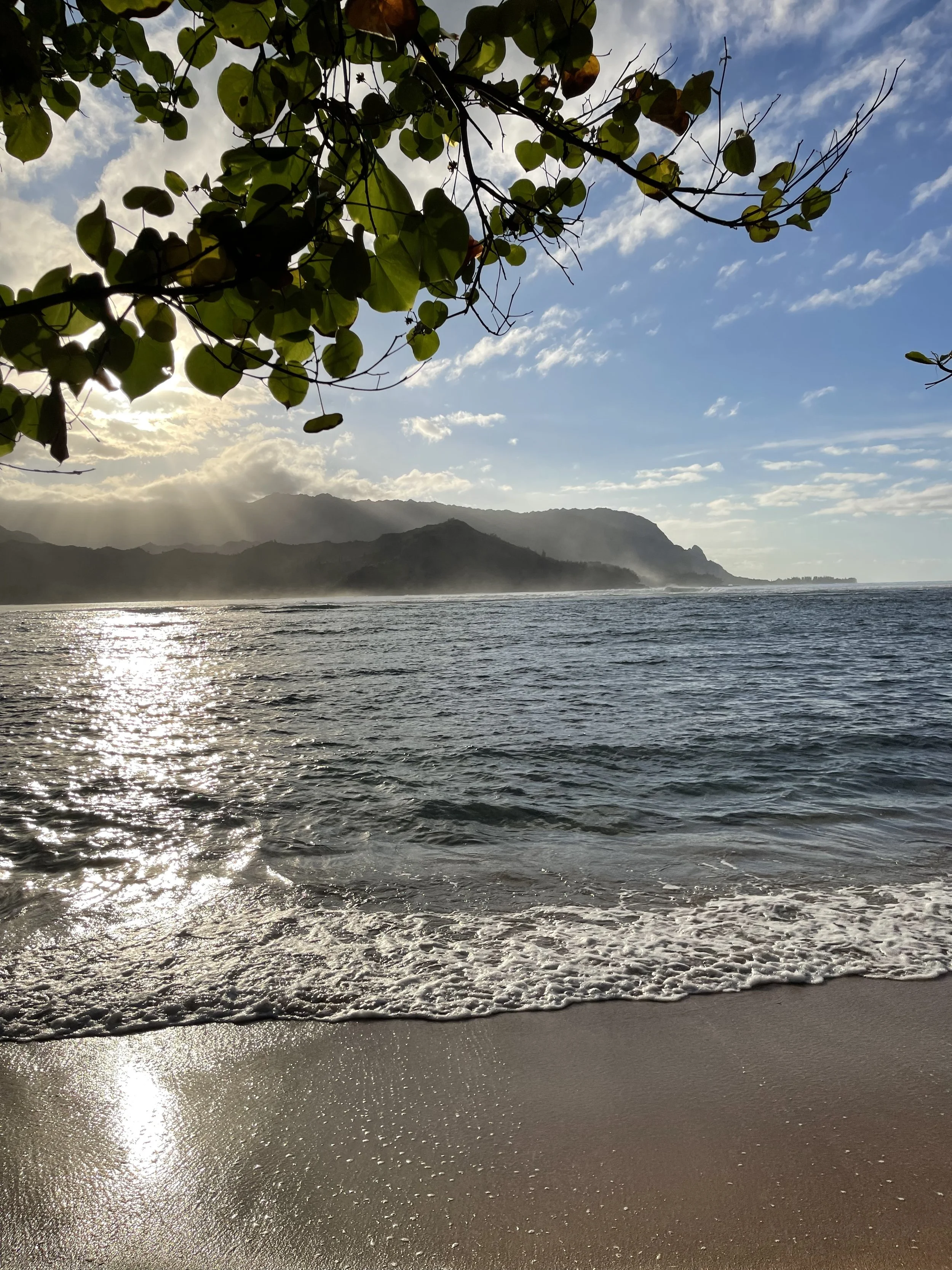 Beach with waves gently rolling onto the sandy shore, overlooking a distant mountainous coastline under a partly cloudy sky with the sun low on the horizon.