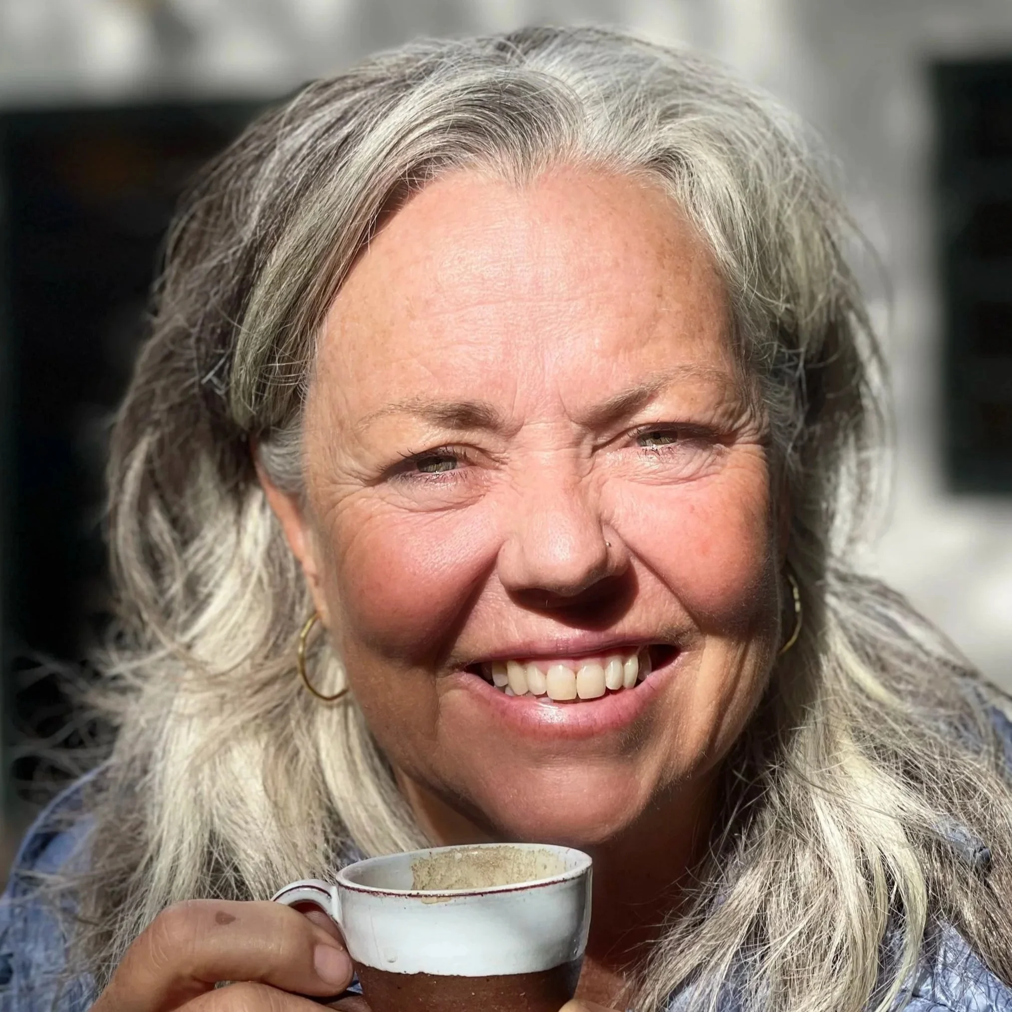 A smiling woman with gray hair holding a cup of coffee with both hands, in front of her face, in a close-up shot.