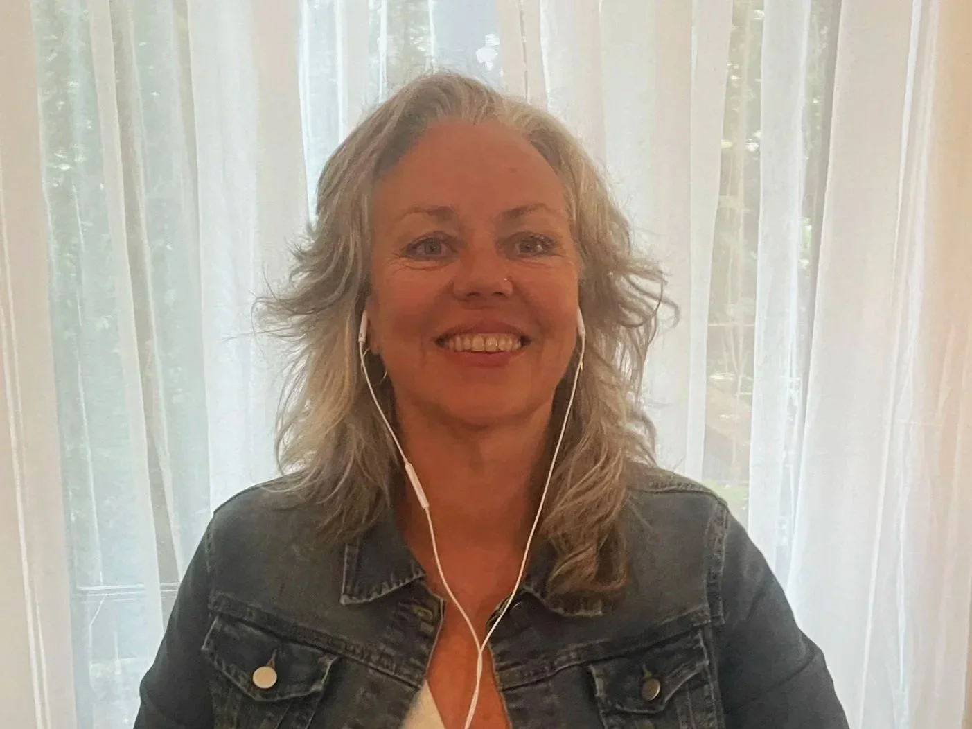 A smiling woman with gray curly hair, wearing a denim jacket and white shirt, with earphones, sitting in front of a sheer white curtain.