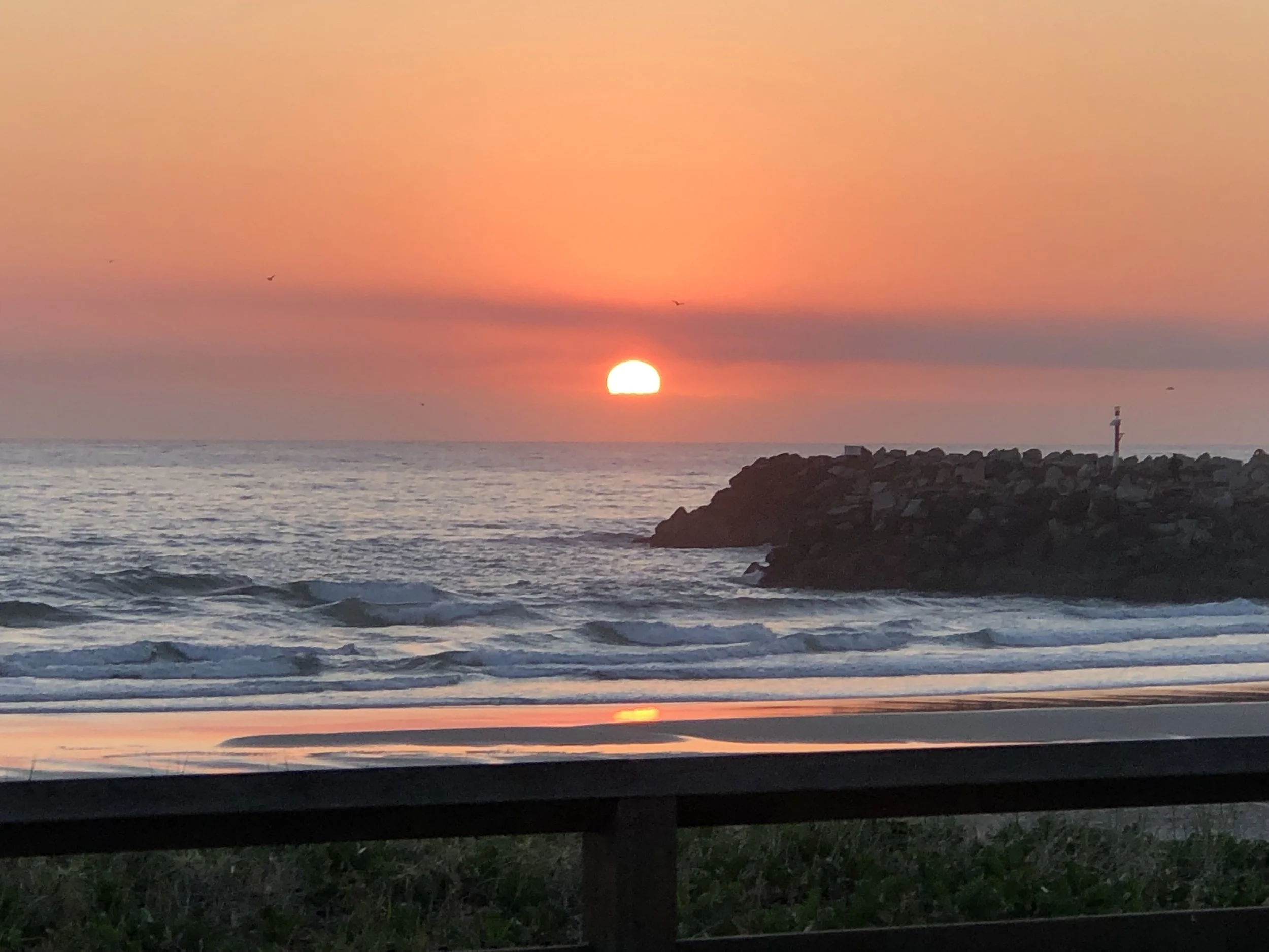Sunset over the ocean with pink and orange sky, a few birds flying, rocky jetty or breakwater, and a lighthouse in the distance.