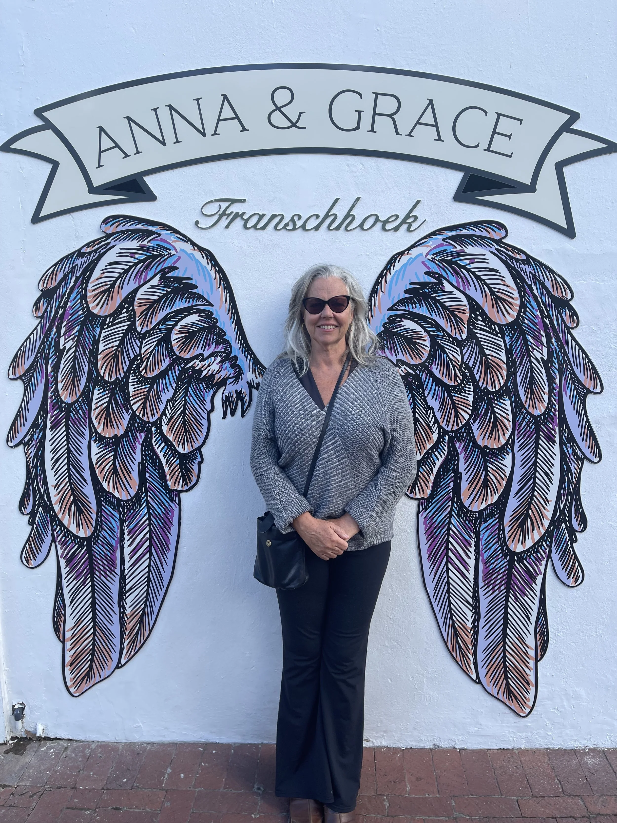 A woman with gray hair and sunglasses standing in front of a wall with colorful angel wings artwork. The wall also has a banner with the text 'ANNA & GRACE' and 'Franschhoek' below it.