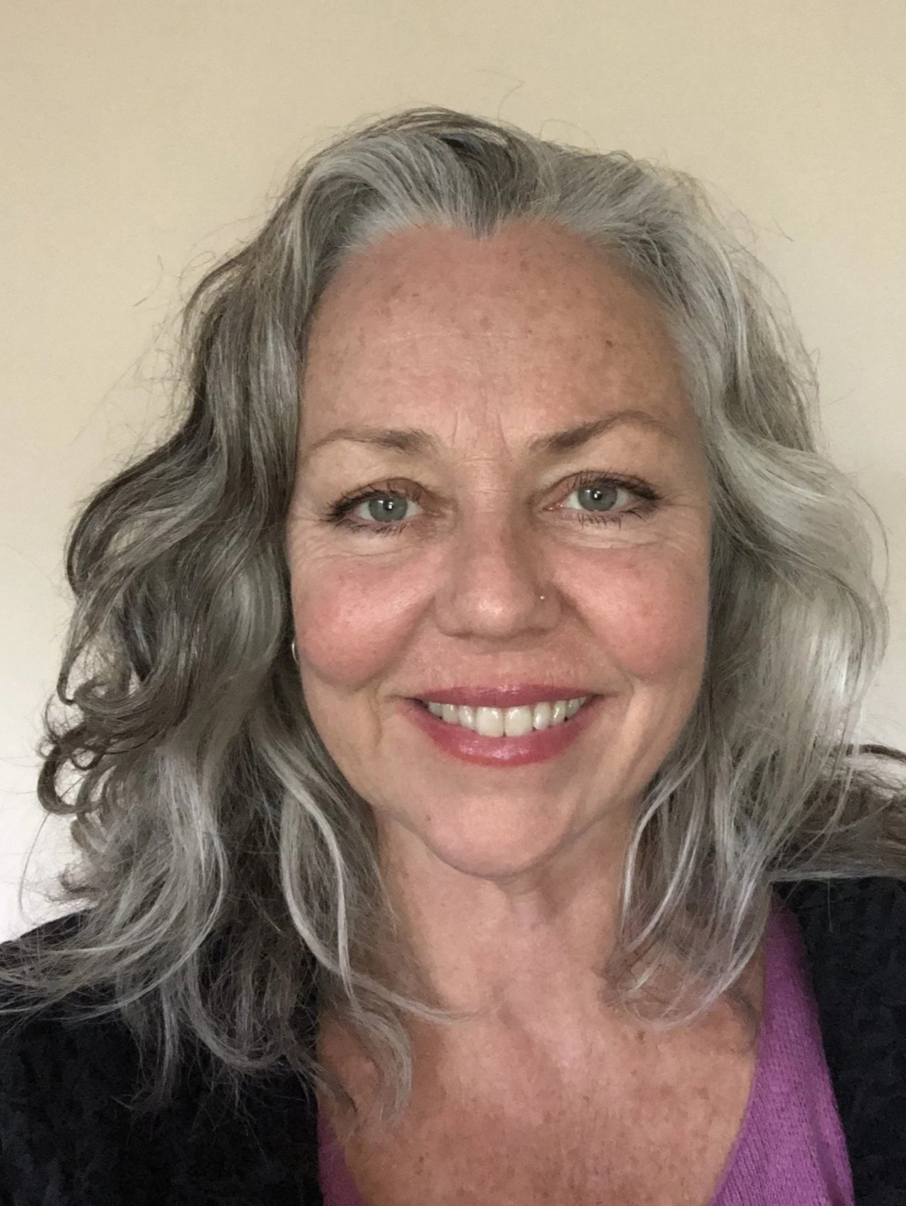 A smiling woman with curly gray hair and green eyes, wearing a purple top and black jacket, standing against a plain beige background.