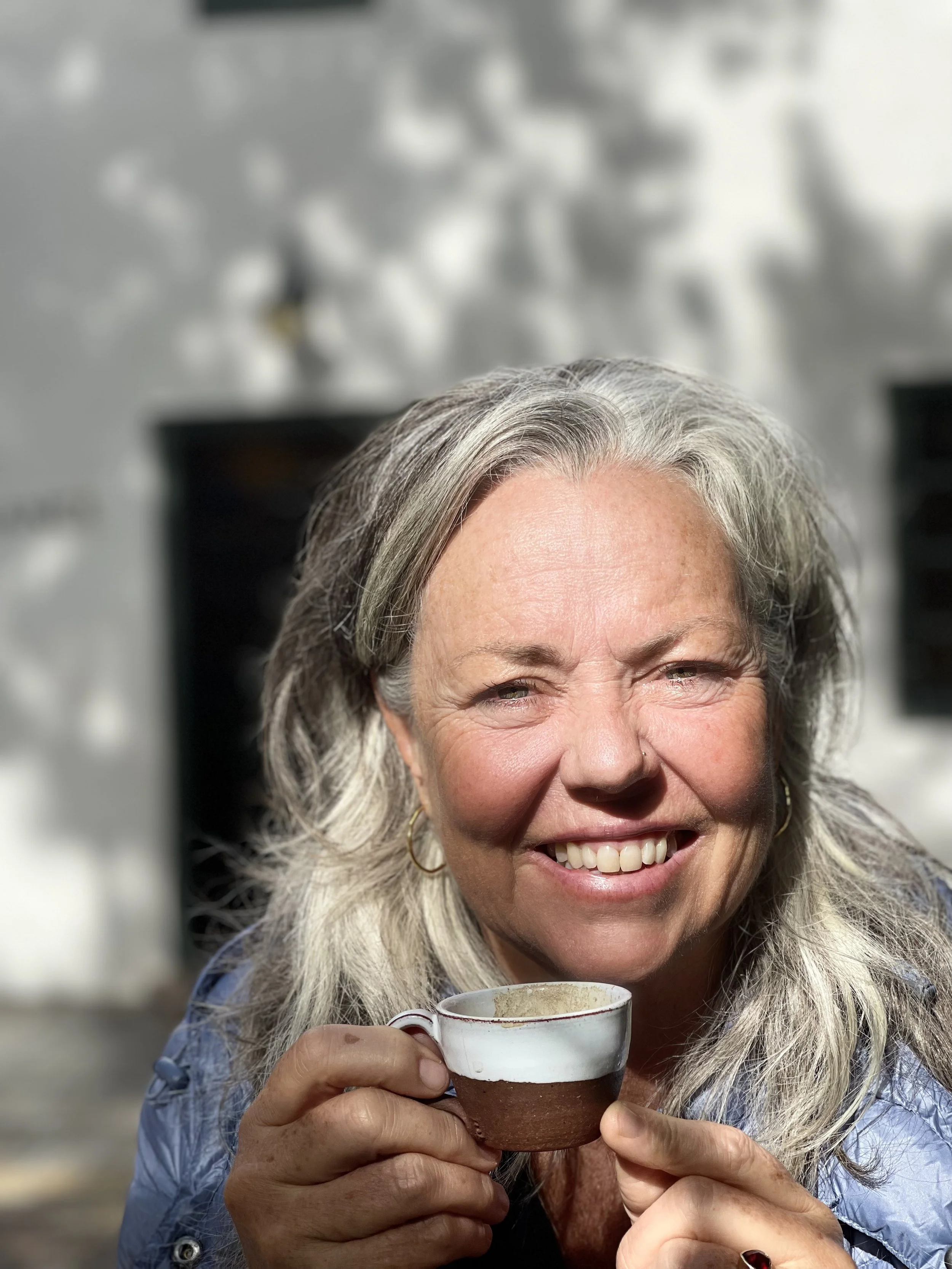 A smiling older woman with gray hair holding a ceramic cup with a red rim, enjoying a warm beverage outdoors.