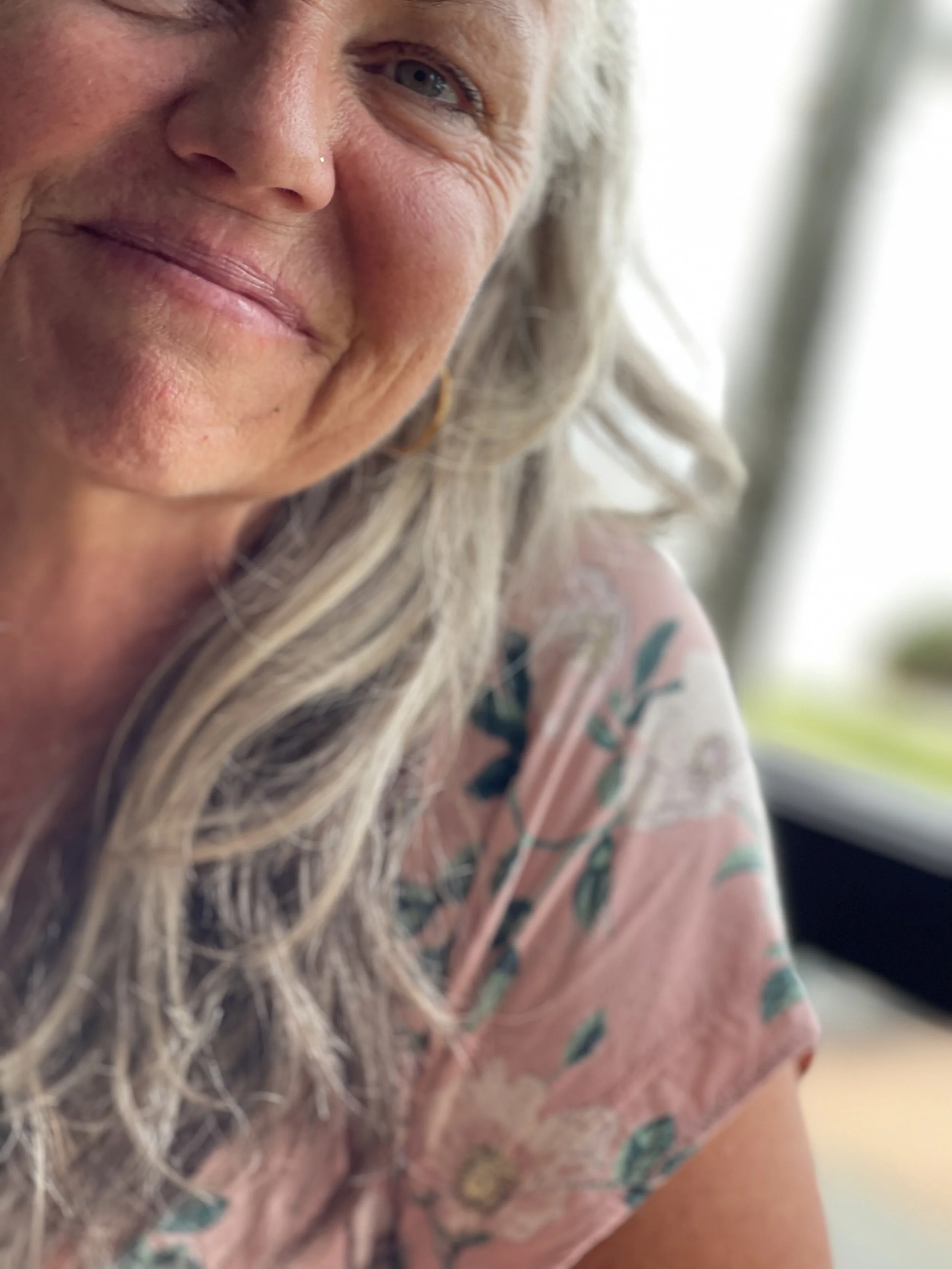 Close-up of a smiling woman with long blonde hair, wearing a pink floral patterned shirt, near a window with natural light.