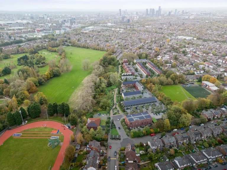 Aerial view of a park with green fields and trees, surrounded by a residential neighborhood with houses, streets, and cars, with a city skyline in the distance.