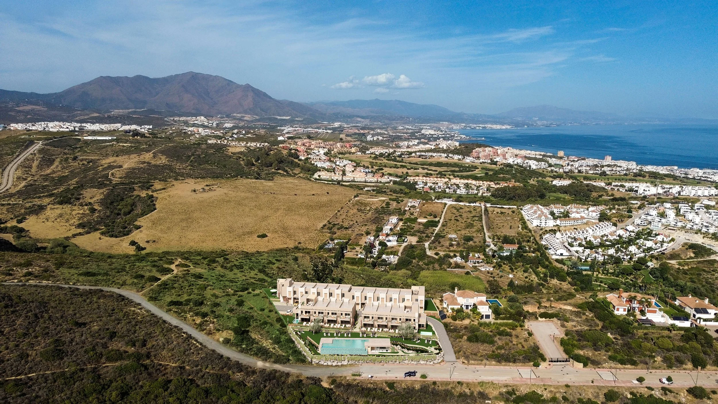 Aerial view of a coastal area with mountains in the background, buildings, houses, and green land, along with a view of the ocean to the right.