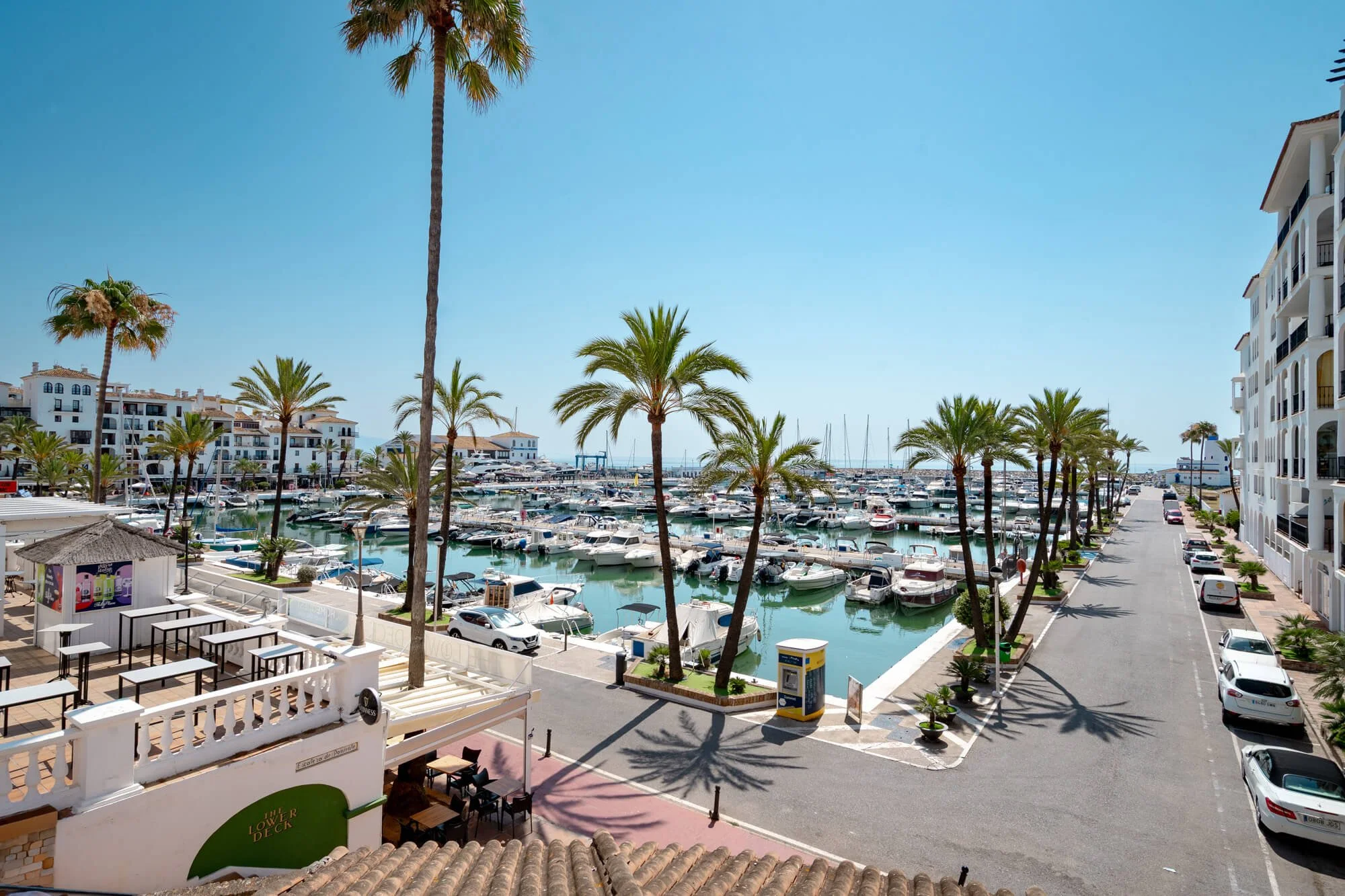 A marina with numerous boats docked, surrounded by palm trees and white multi-story buildings in a sunny, clear sky.
