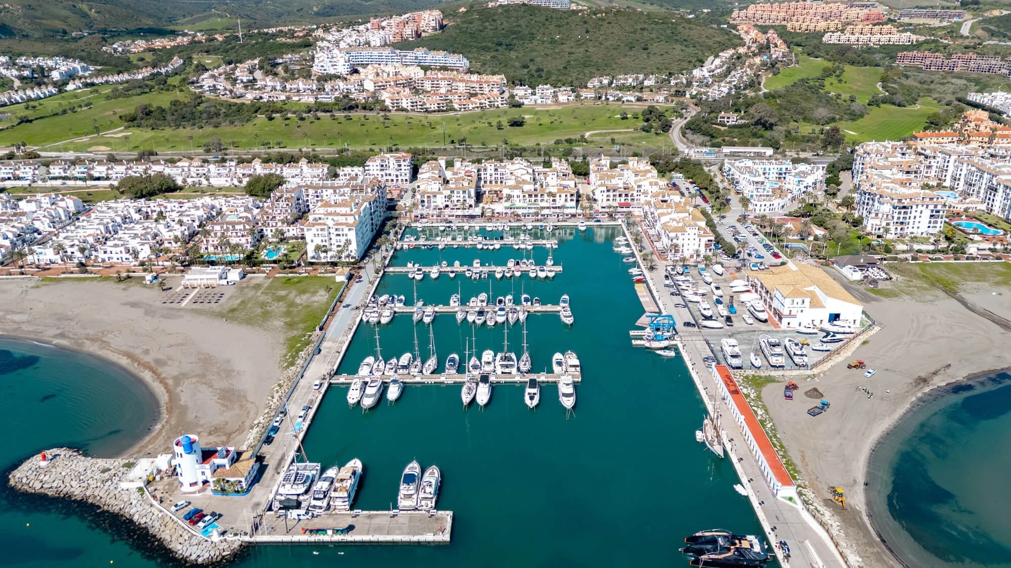 Aerial view of a marina with numerous boats docked, surrounded by white residential buildings, green hills in the background, and sandy beach areas.