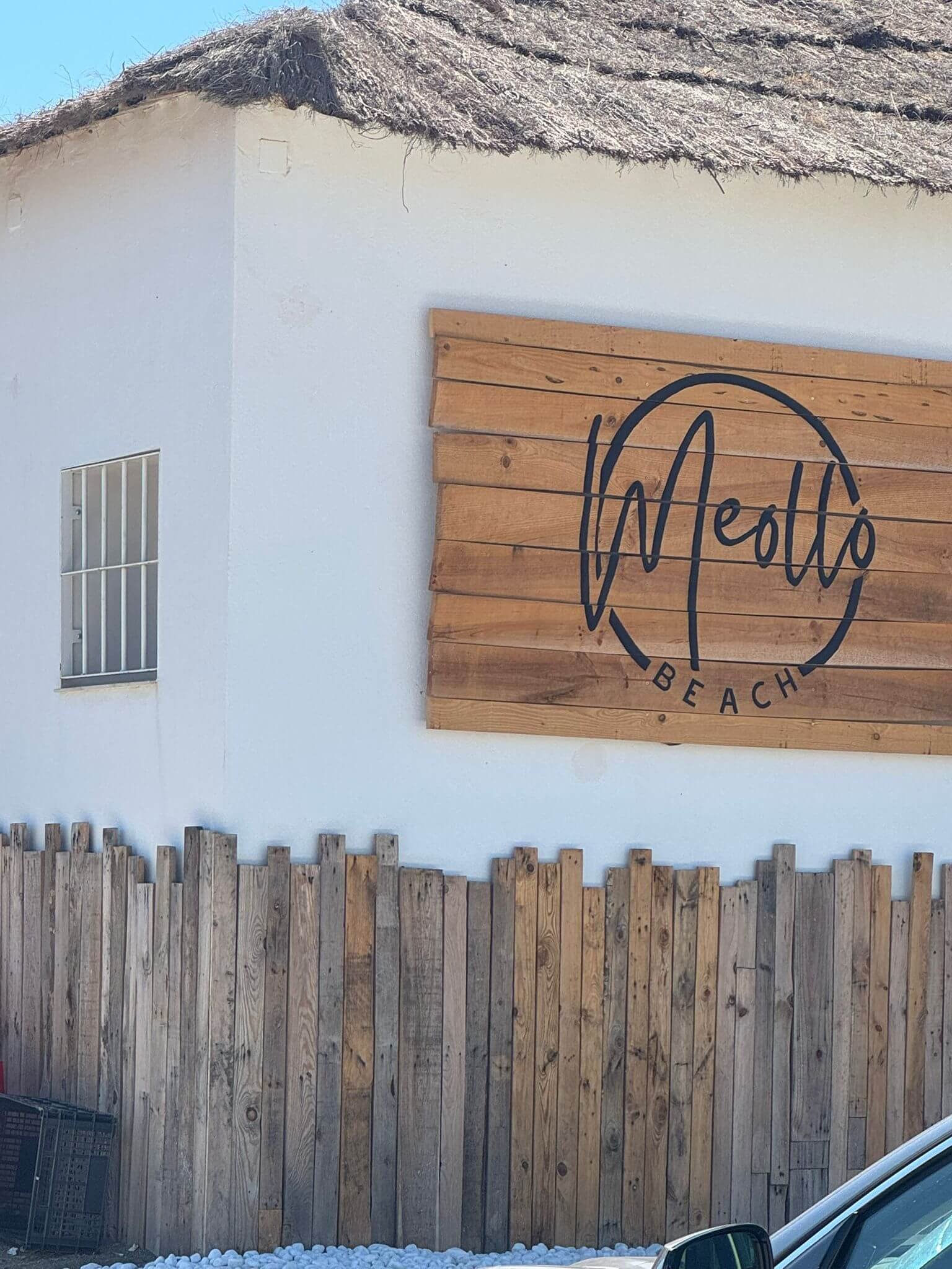 A white building with a thatched roof, a small window with bars, and a large wooden sign reading 'Ollie' with the word 'BEACH' underneath, in front of a wooden fence and some gravel.