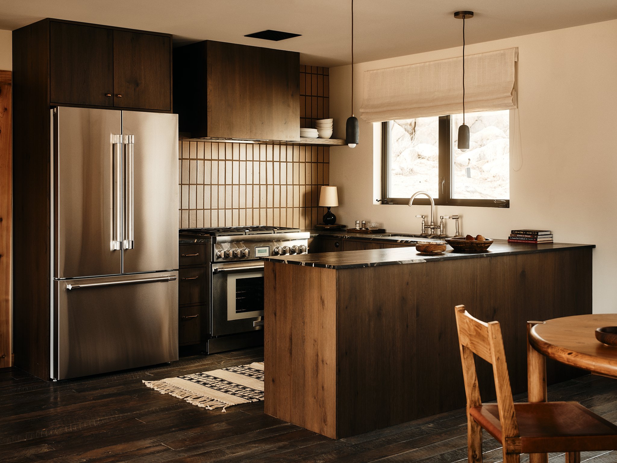 A modern kitchen with dark wood cabinets, stainless steel refrigerator, oven, and stovetop, and a window above the sink. There's a small table with wooden chairs and minimal decor, including a lamp and a stack of books.