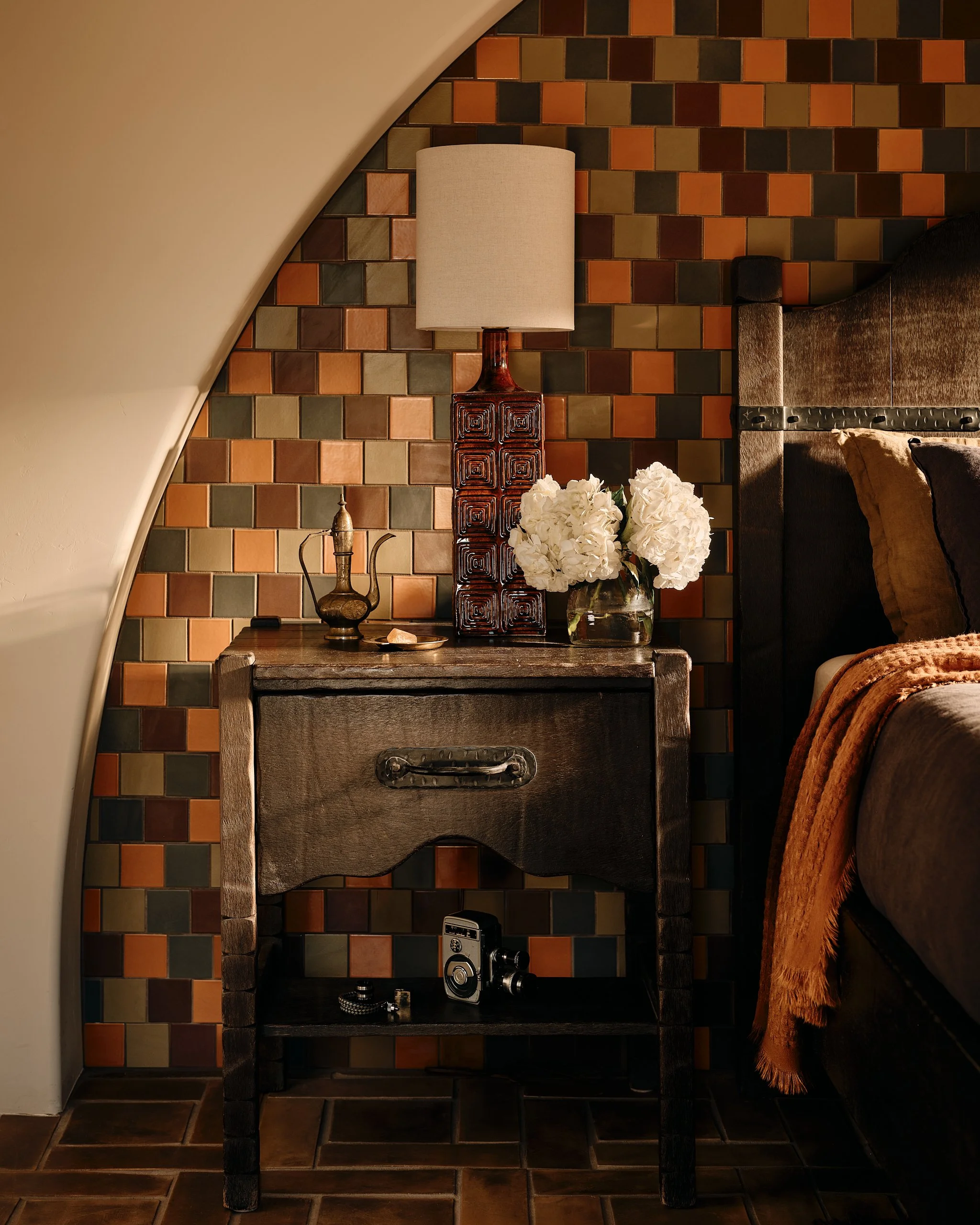 Close-up of a dark wood bedside table against a multi-colored tiled wall. The table has a vintage camera, a small tray with items, a vase with white flowers, a tall table lamp with a beige shade, on top. Part of a bed with brown and black pillows and