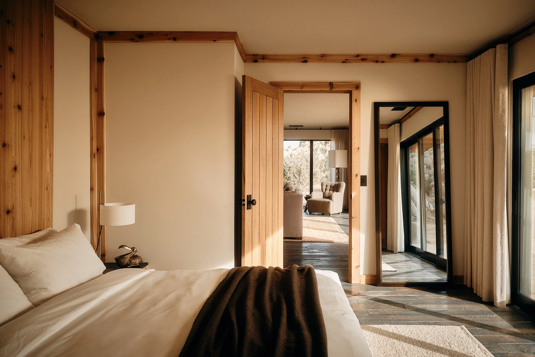 Interior view of a bedroom with a wooden headboard, white bedding, a bedside lamp, and a view of a living room through an open door. The room has wood trim and a sliding glass door with curtains leading outside.
