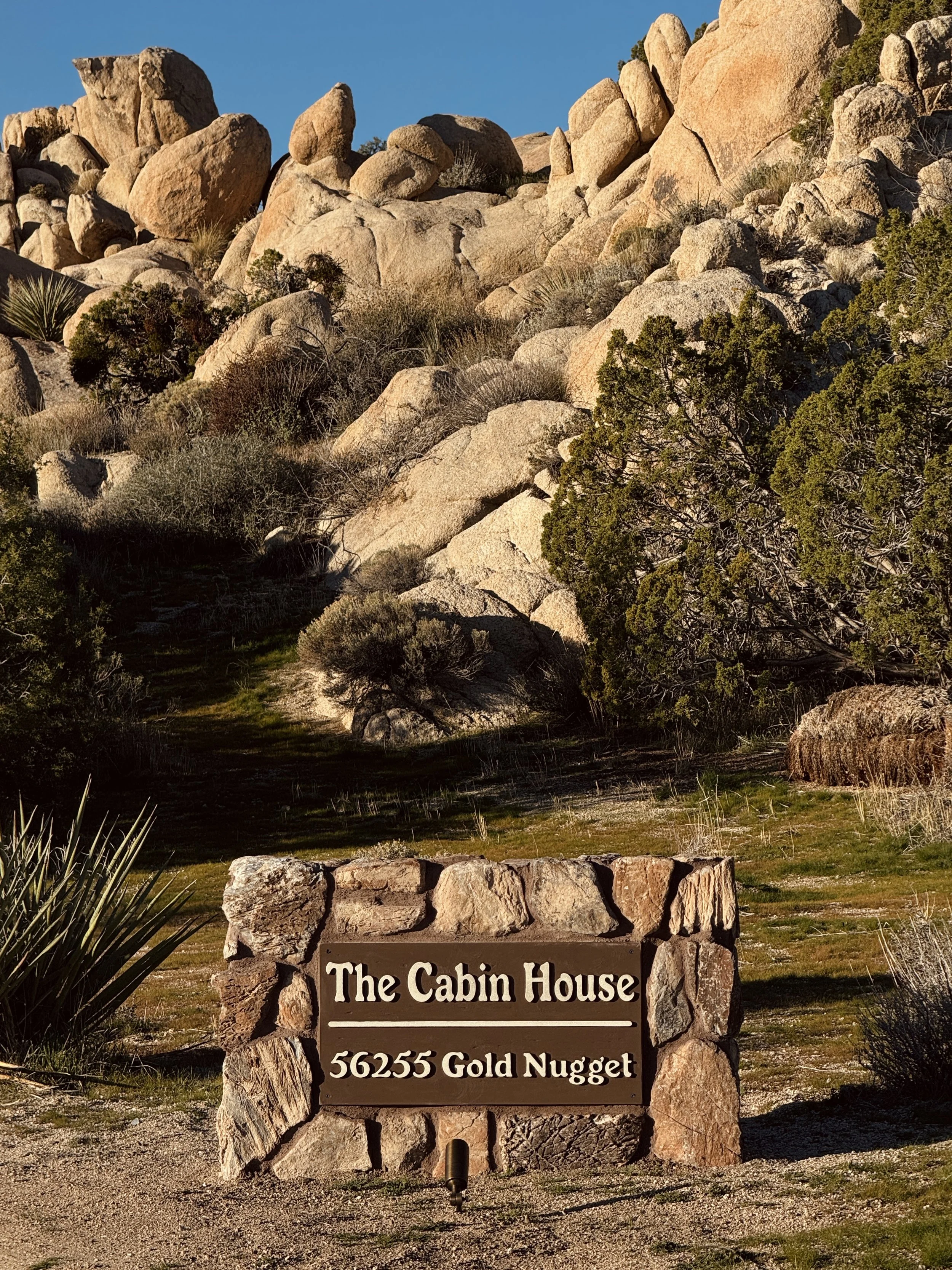 A stone sign in front of a desert landscape with large rocks and sparse vegetation, displaying the text "The Cabin House, 56255 Gold Nugget."