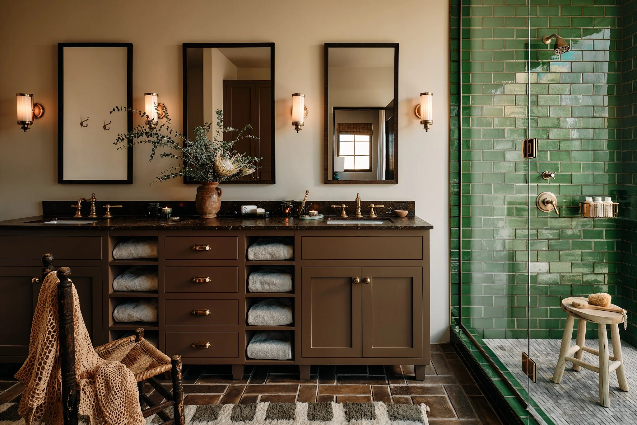 A bathroom featuring a double vanity with a dark countertop, three framed mirrors, and wall-mounted lights. There are potted plants and toiletries on the counter. To the right, there is a green-tiled walk-in shower with a glass door, a wooden stool w
