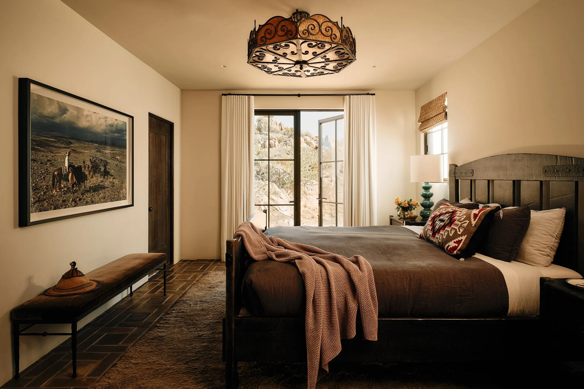 A cozy bedroom with a black wooden bed adorned with decorative pillows and a brown duvet, a white lamp with a turquoise base, a small bouquet of flowers, a large painting of horses and a person on horseback, a bench with a hat, and a large window wit