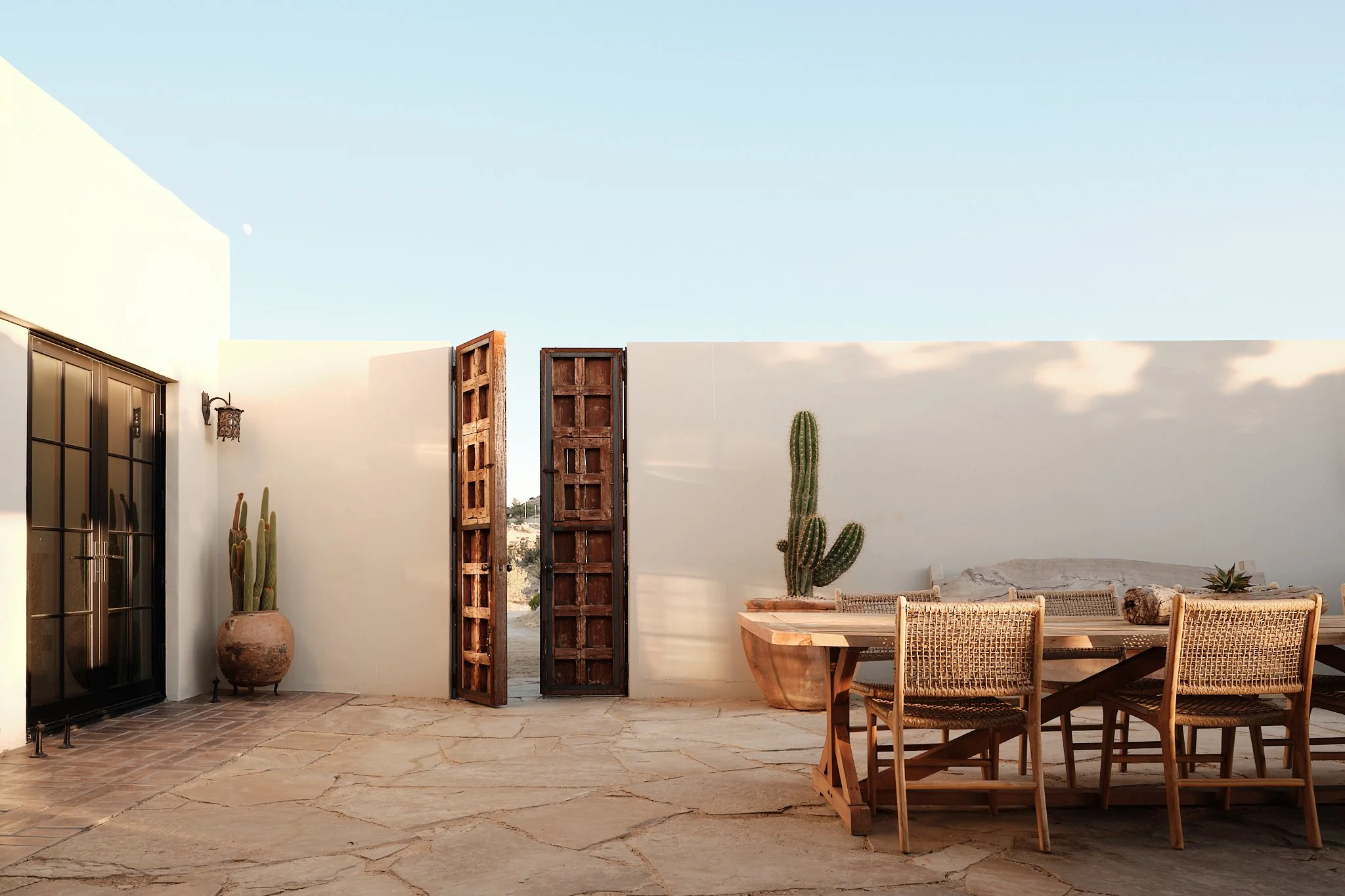 An outdoor patio with a stone floor, wooden furniture, and potted cacti, enclosed by a white wall and open wooden gates leading to sand and distant terrain, under a clear sky.