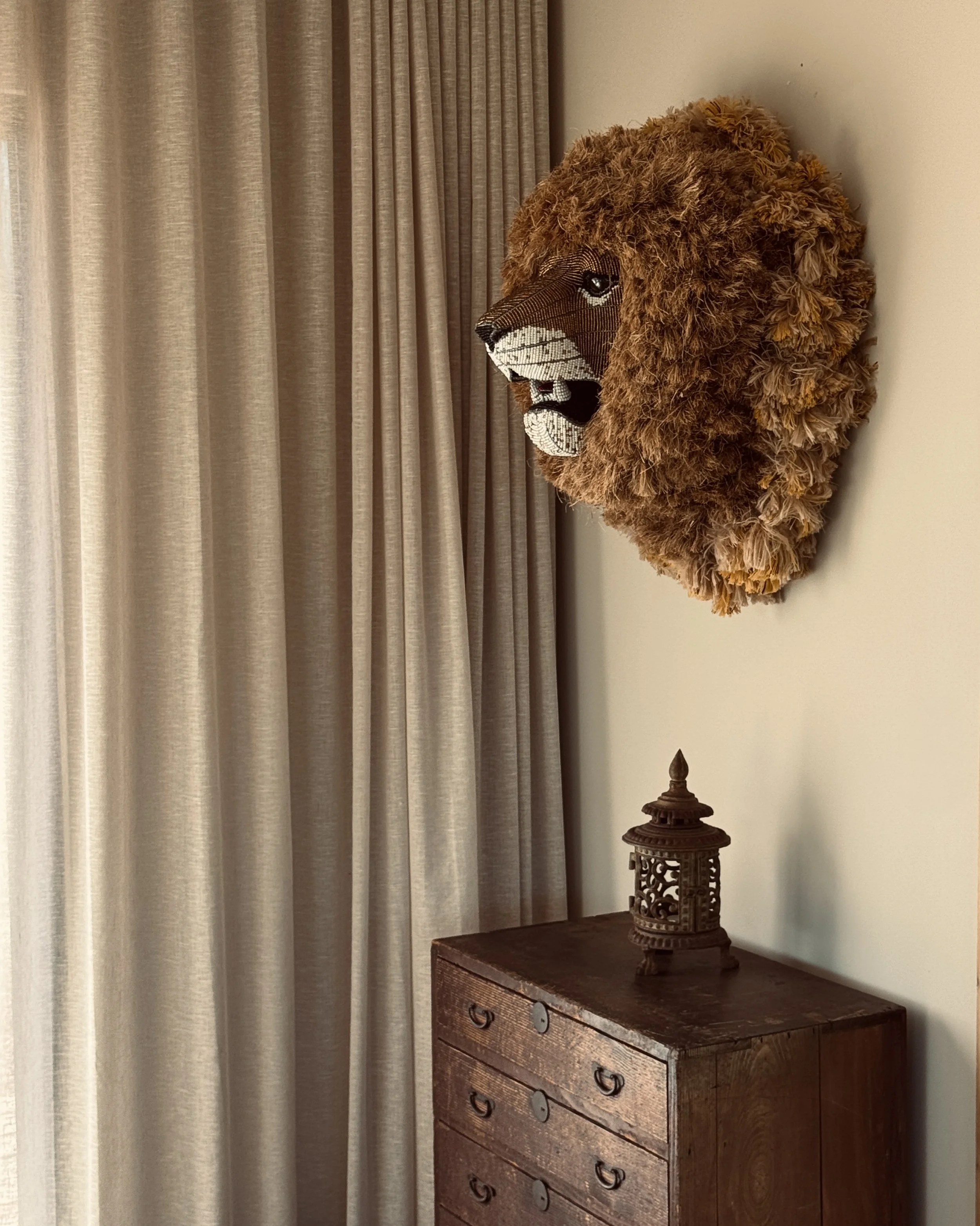 A lion's head wall art with a mane made of textured fabric hanging above a wooden dresser with a small decorative lantern on top, next to beige curtains.