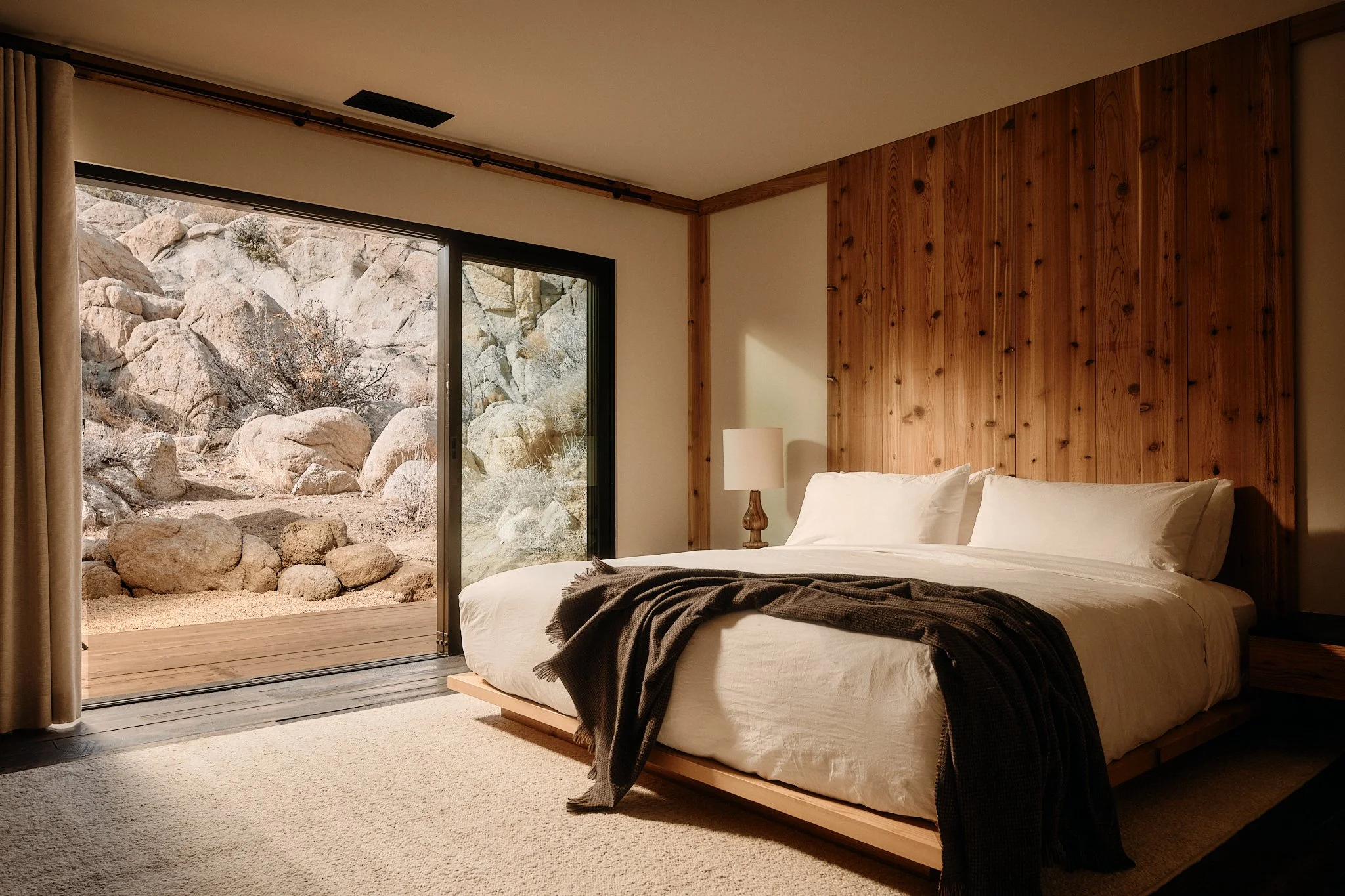 A bedroom with a large bed, wooden headboard, and a nightstand with a lamp. Sliding glass door opens to a rocky outdoor landscape.