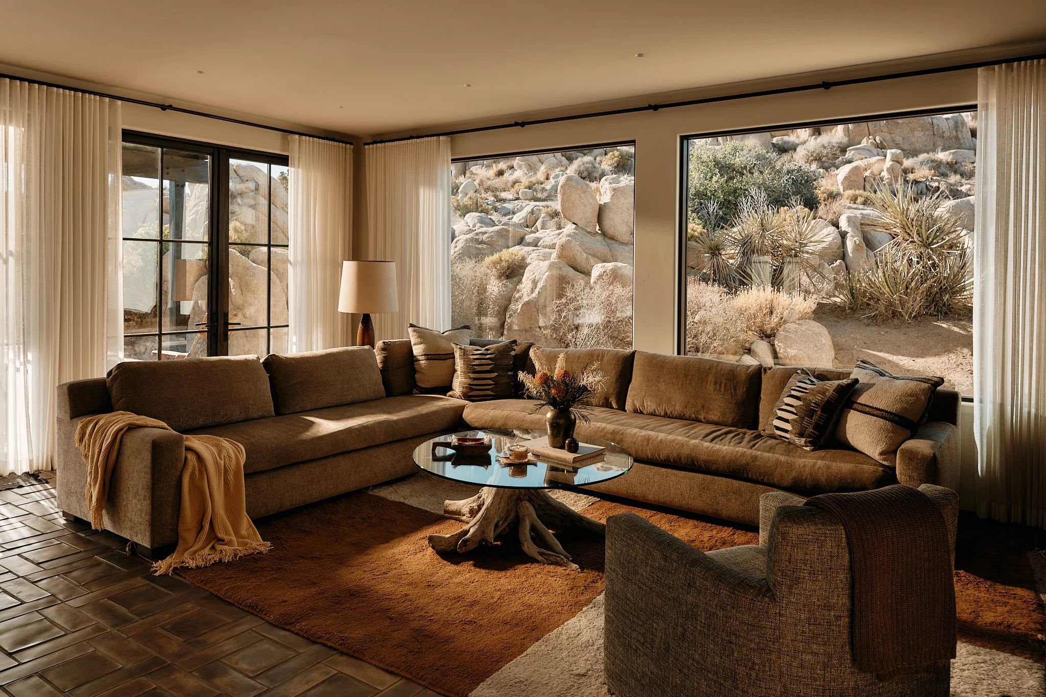 A swimming pool outdoors in a desert landscape with rocks, trees, lounge chairs, umbrellas, and a woven sitting area.