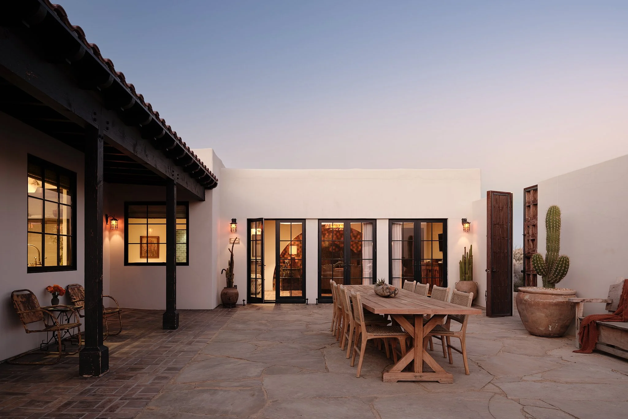 An outdoor patio area with a large wooden dining table surrounded by chairs, desert plants in pots, and a building with large windows and glass doors, during sunset.