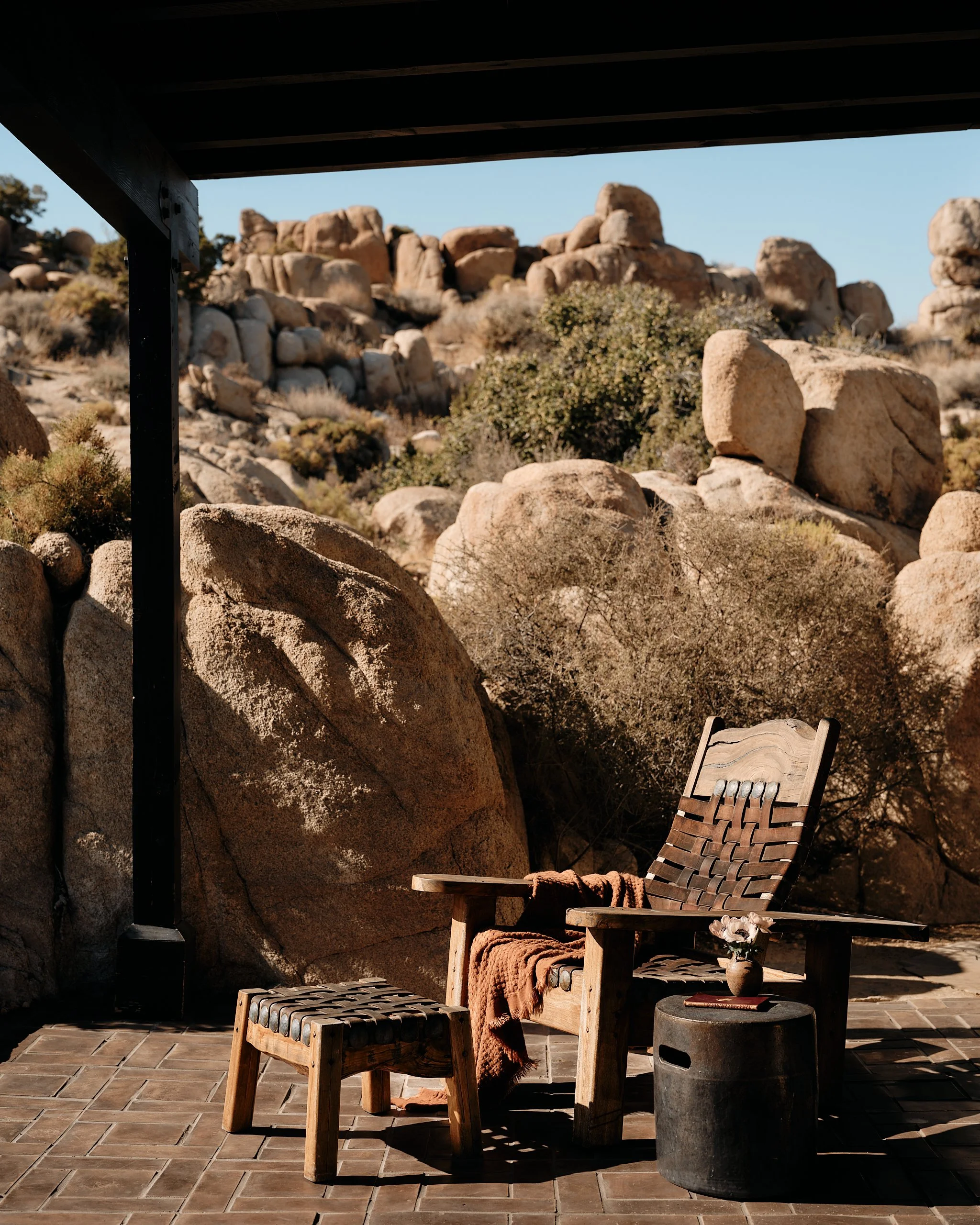 A wooden armchair with a blanket draped over it, a small stool, and a flower in a vase on top of a cylindrical table, set on a patio in a rocky desert landscape with large boulders and dry bushes.