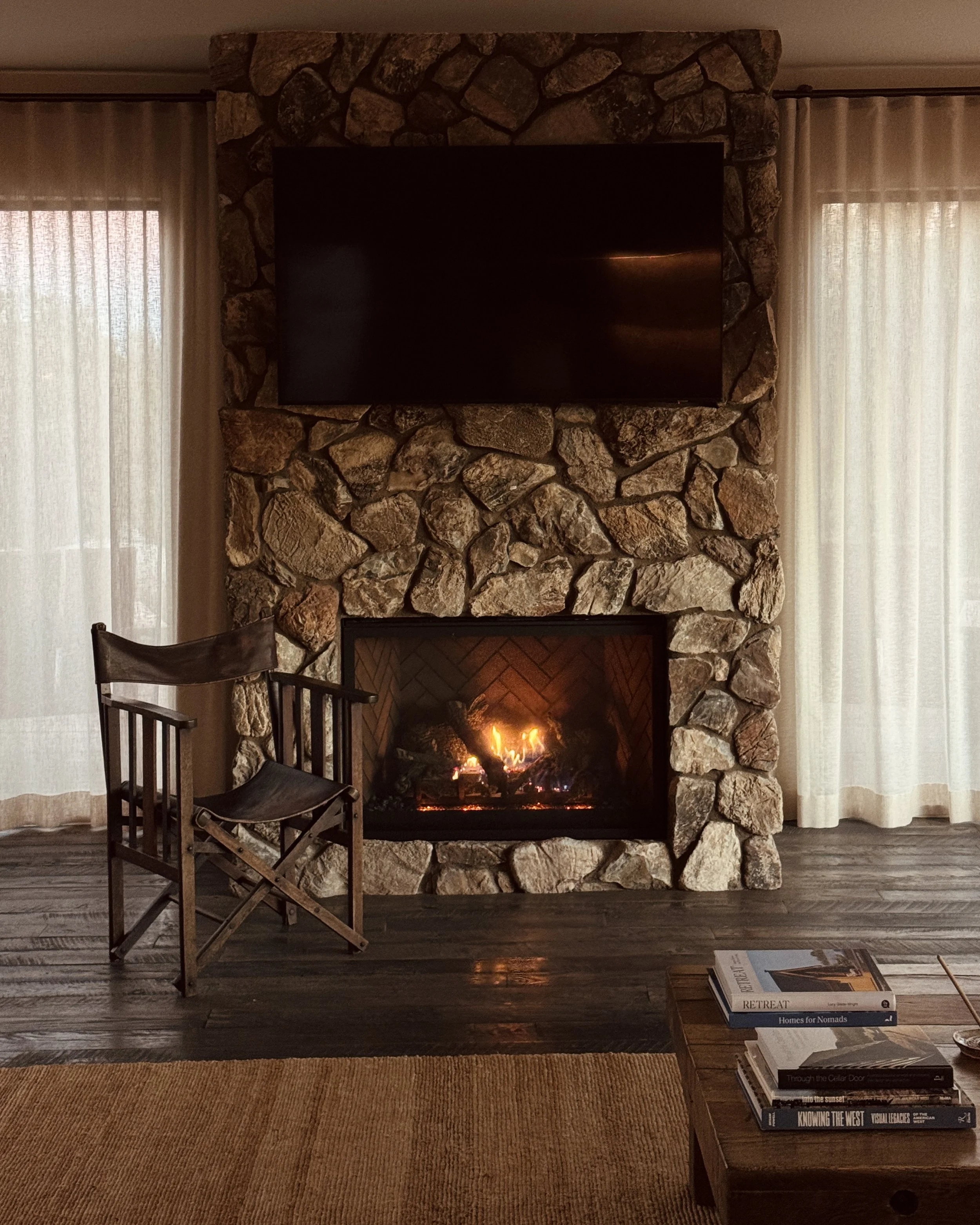 A cozy living room with a stone fireplace, a mounted flat-screen TV above, a wooden chair to the left of the fire, sheer curtains on both sides, and a coffee table with books in the foreground.