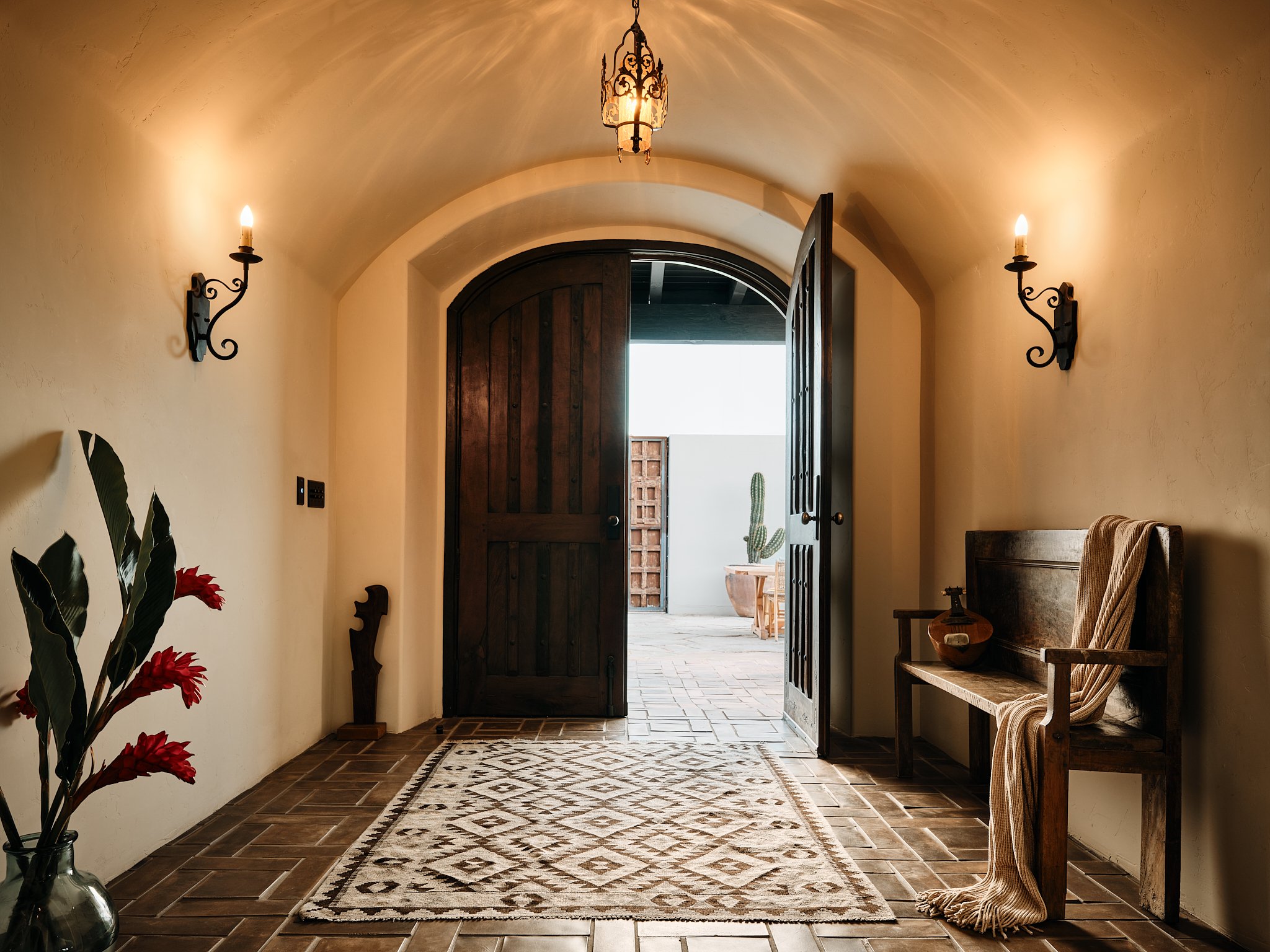 Entrance hallway with open wooden door leading outside, potted cactus, bench with a blanket, decorative vase, and a patterned rug on tile flooring, illuminated by warm wall-mounted lights and a hanging ceiling light.