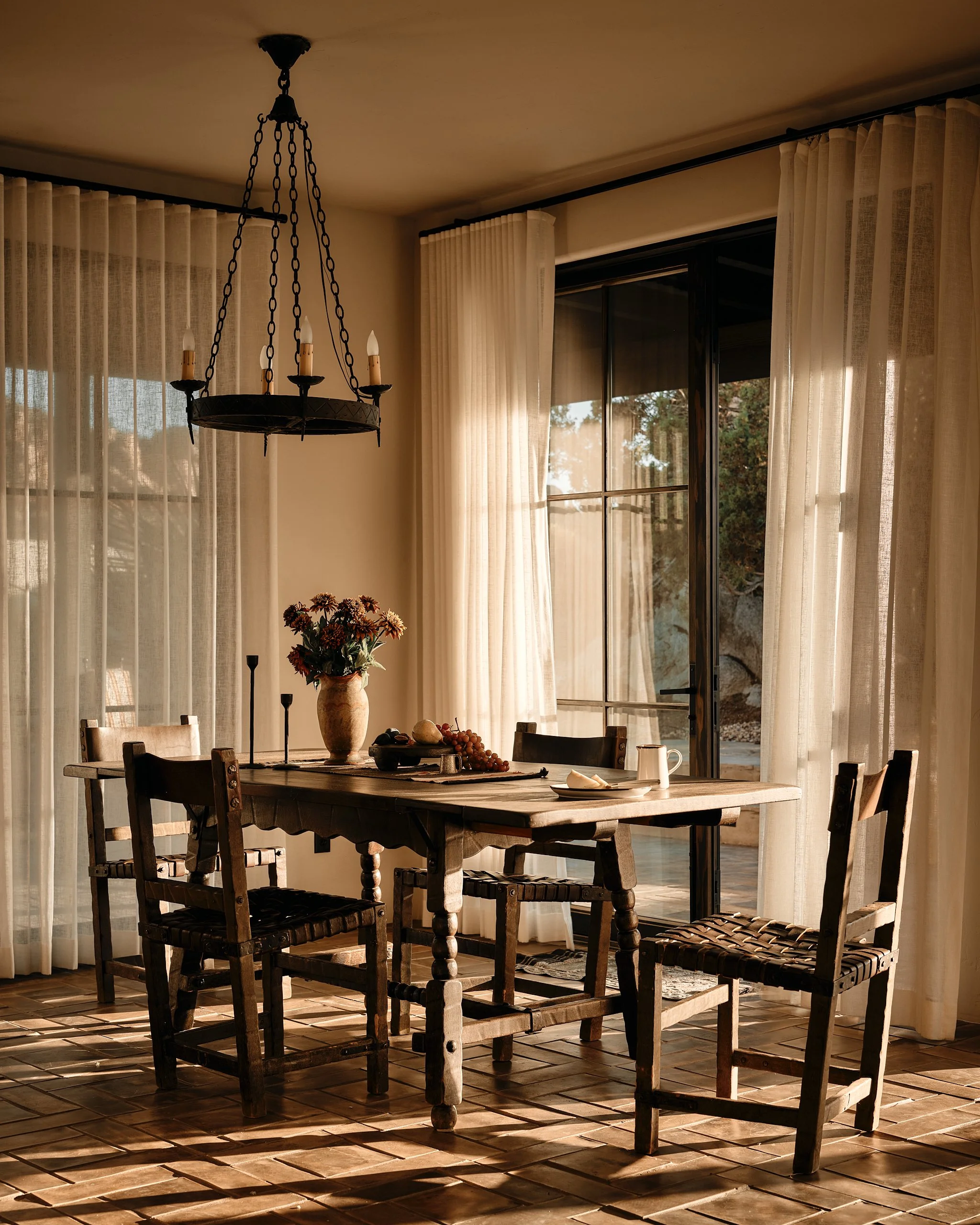 A rustic dining room with a wooden table set with a vase of flowers, fruit, and a pitcher, surrounded by four wooden chairs, illuminated by natural sunlight coming through large windows with white curtains, and a black metal chandelier hanging from t