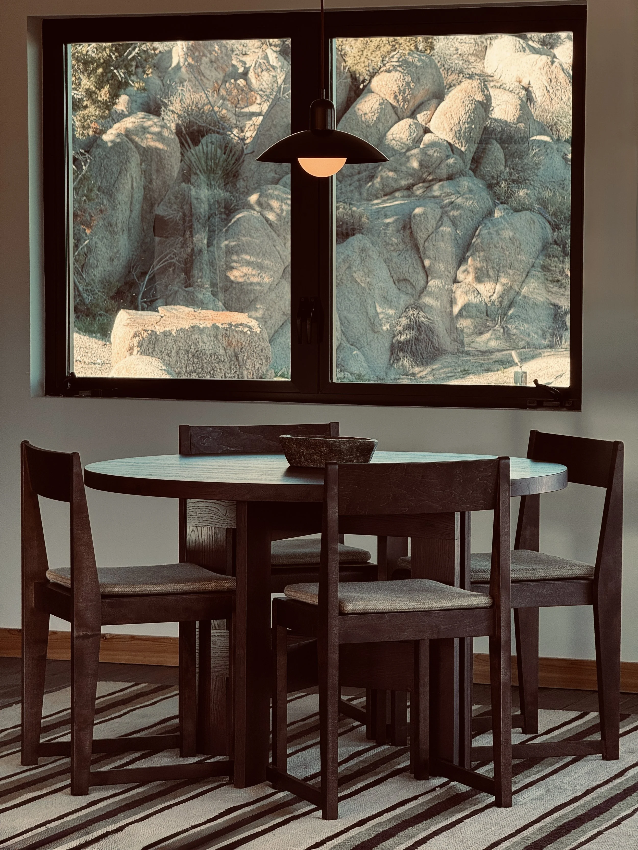 A round wooden dining table with four wooden chairs with cushioned seats, a bowl on the table, and a large window showing a rocky desert landscape outside, with a hanging pendant lamp above the table.