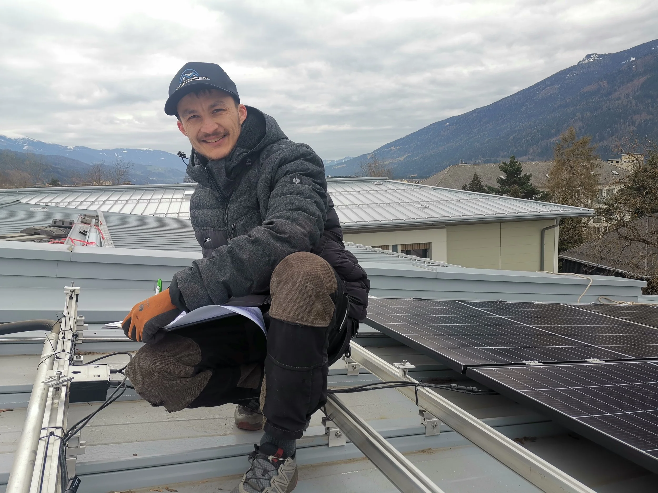 Ein Mann auf einem Dach mit Solarpanelen unter einem bewölkten Himmel, umgeben von Bergen und Häusern.
