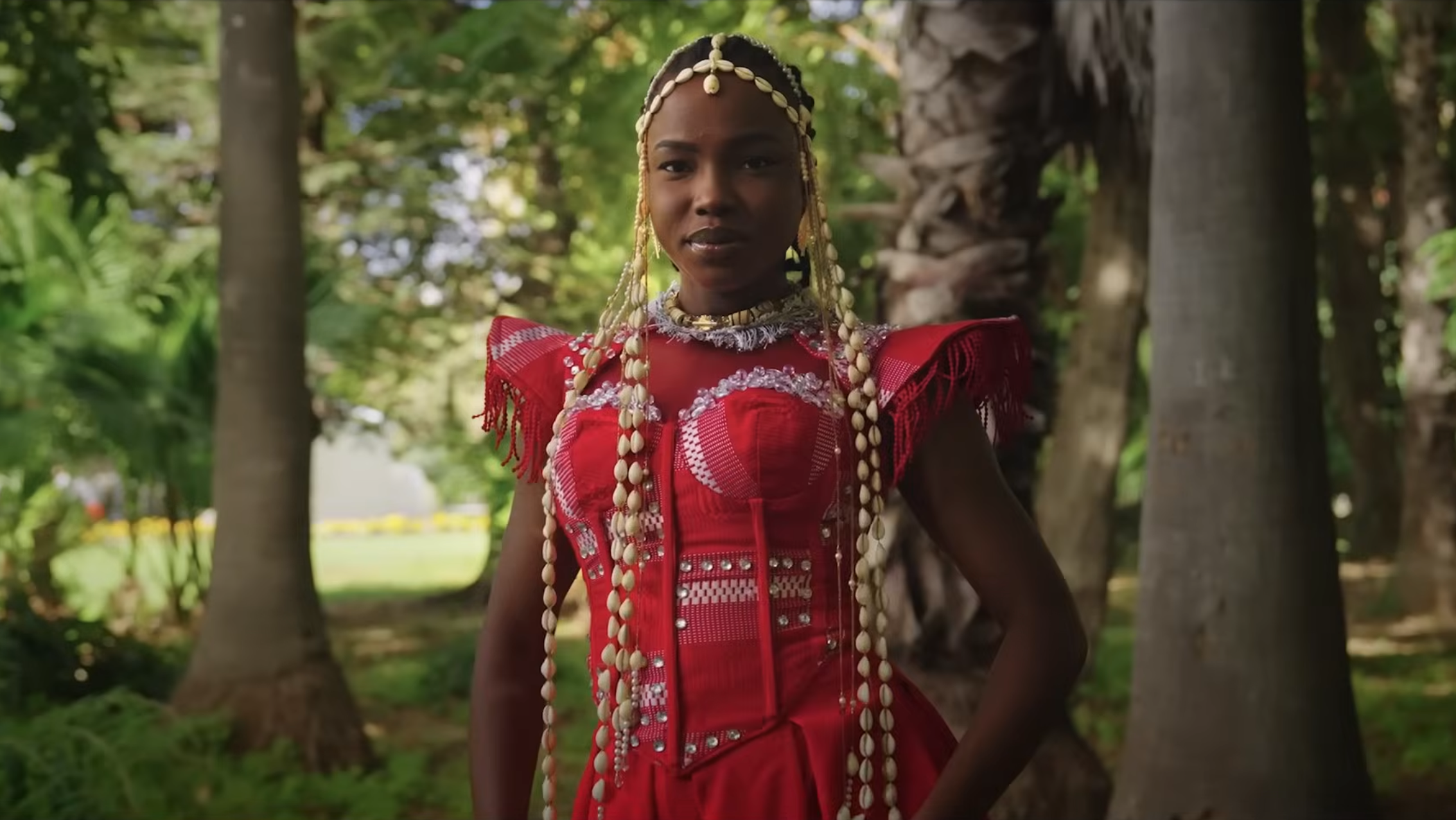 A woman dressed in traditional African attire standing outdoors near trees with green foliage.