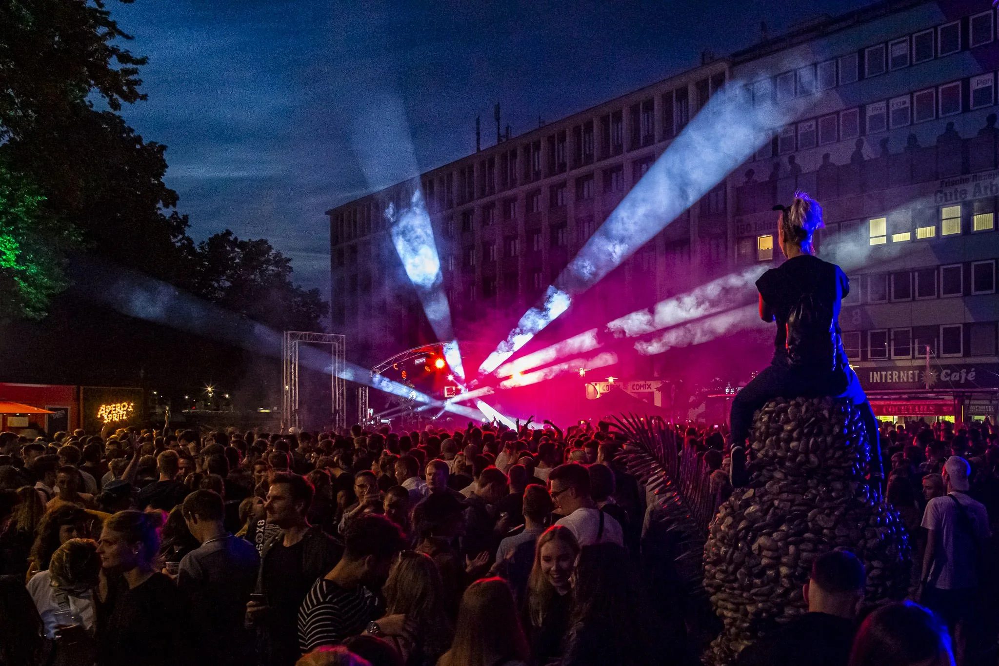Eine Menschenmenge schaut sich ein nächtliches Open-Air-Konzert an, bei dem bunte Scheinwerferstrahlen auf die Bühne gerichtet sind, im Hintergrund ein mehrstöckiges Gebäude mit gekachelter Fassade.