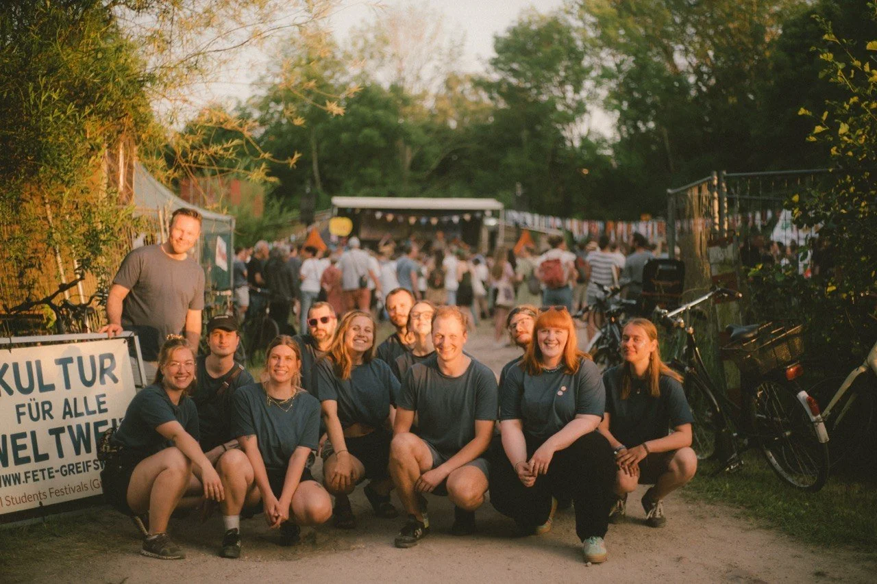 Gruppenfoto des ehrenamtlichen Orga-Teams der Fête de la Musique Greifswald 2025 (GrIStuF e.V.) vor einem Banner mit der Aufschrift „Kultur für alle weltweit" auf dem Festivalgelände.