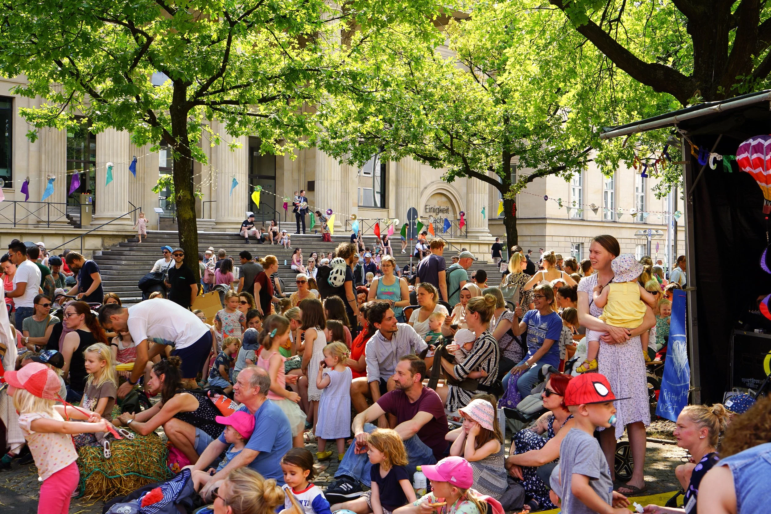 Menschen versammeln sich bei einem öffentlichen Event im Freien, Kita 1, unter Bäumen vor einem Gebäude mit Säulen und einer Inschrift. Kinder, Erwachsene und Familien sitzen und stehen auf den Stufen und im Bereich davor. Es gibt bunte Fahnen und Dekorationen.