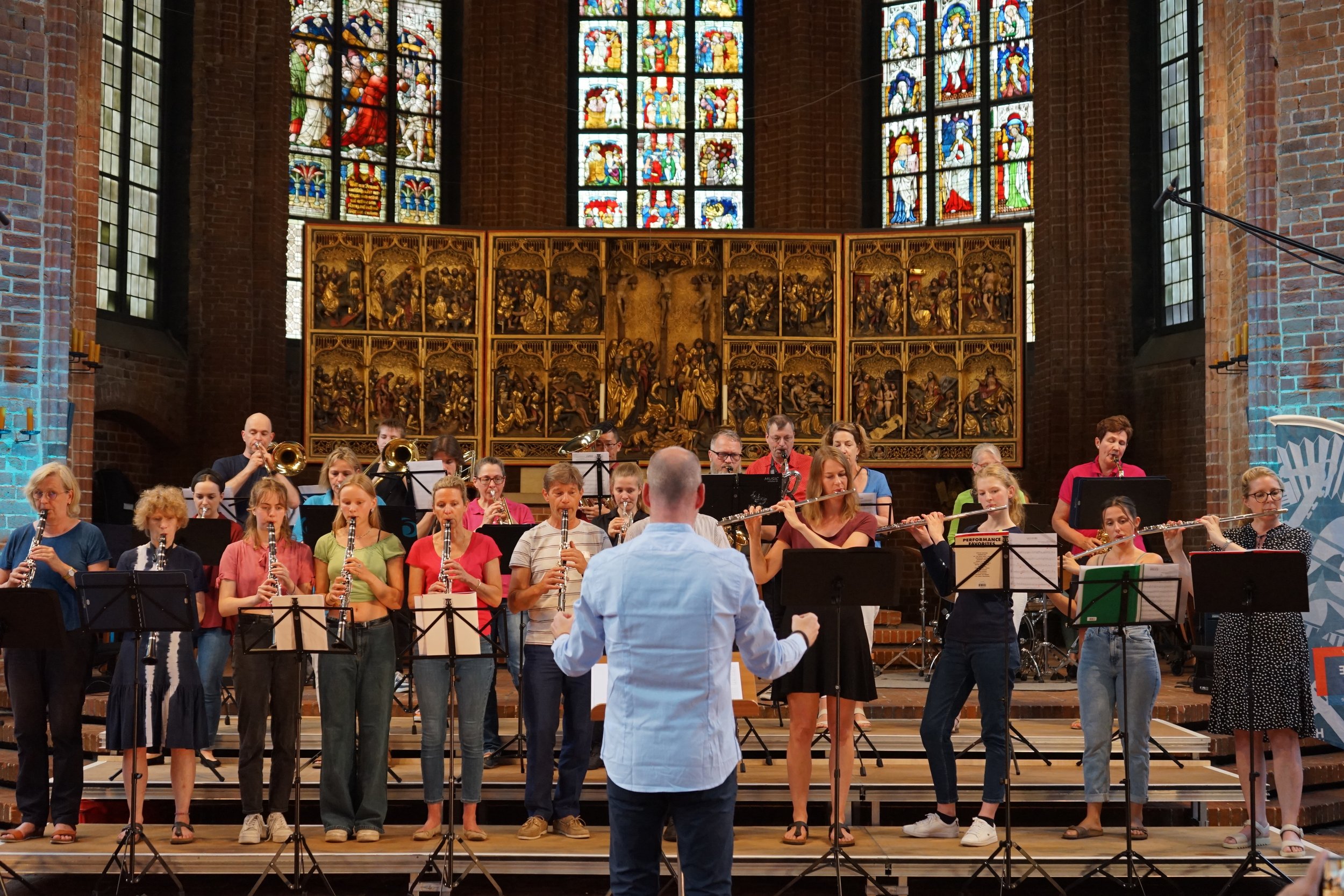 Eine Gruppe von Menschen, darunter Kinder und Erwachsene, die in einer Kirche oder einem Konzertsaal mit Holzpodest und Wandkunst musizieren, während ein Dirigent sie dirigiert. Im Hintergrund sind große bunte Glasfenster und eine kunstvolle Kirchenfensterwand sichtbar.