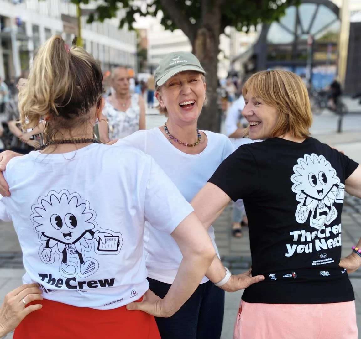 Drei lachende Frauen in einem Kreis, die sich umarmen, im Freien auf einer Straße, tragen T-Shirts mit einem Cartoon-Charakter und dem Text 'The Crew'.
