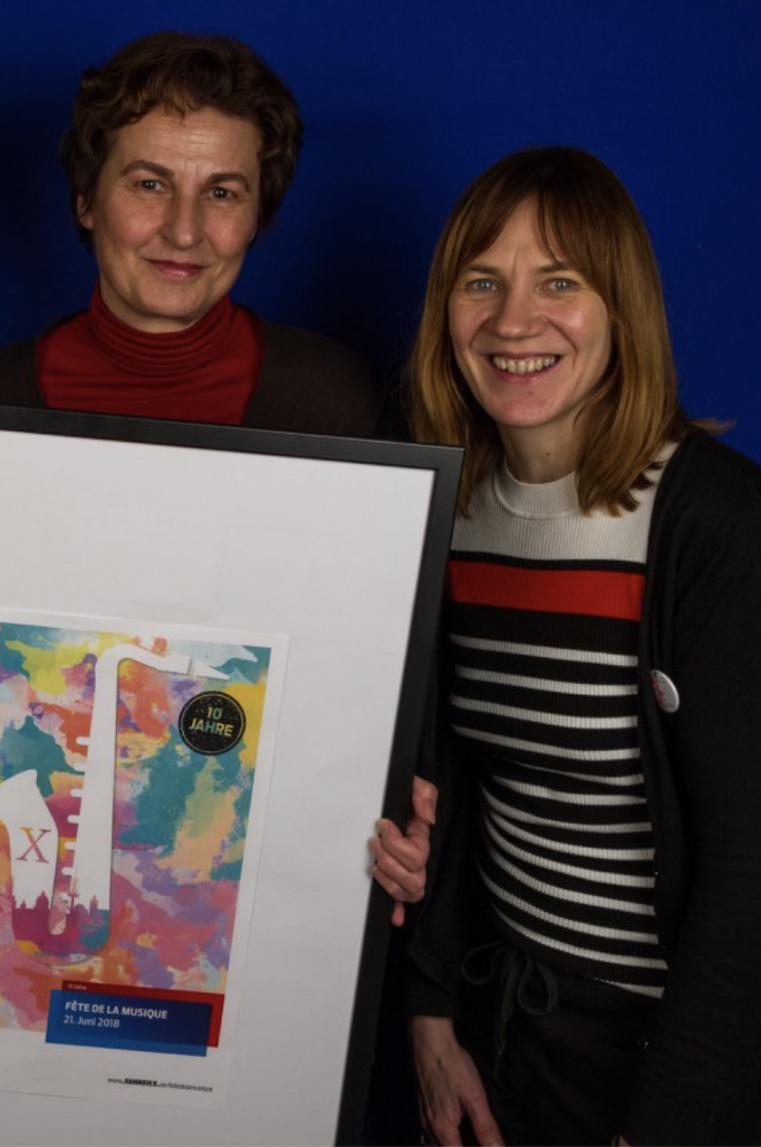 Foto von Simone Hofmann, Gründerin des Fête de la Musique Deutschland Netzwerks, und Sabine Busmann, Geschäftsführerin des MusikZentrum Hannover, mit einem gerahmten Jubiläumsplakat zum 10-jährigen Bestehen der Fête de la Musique.