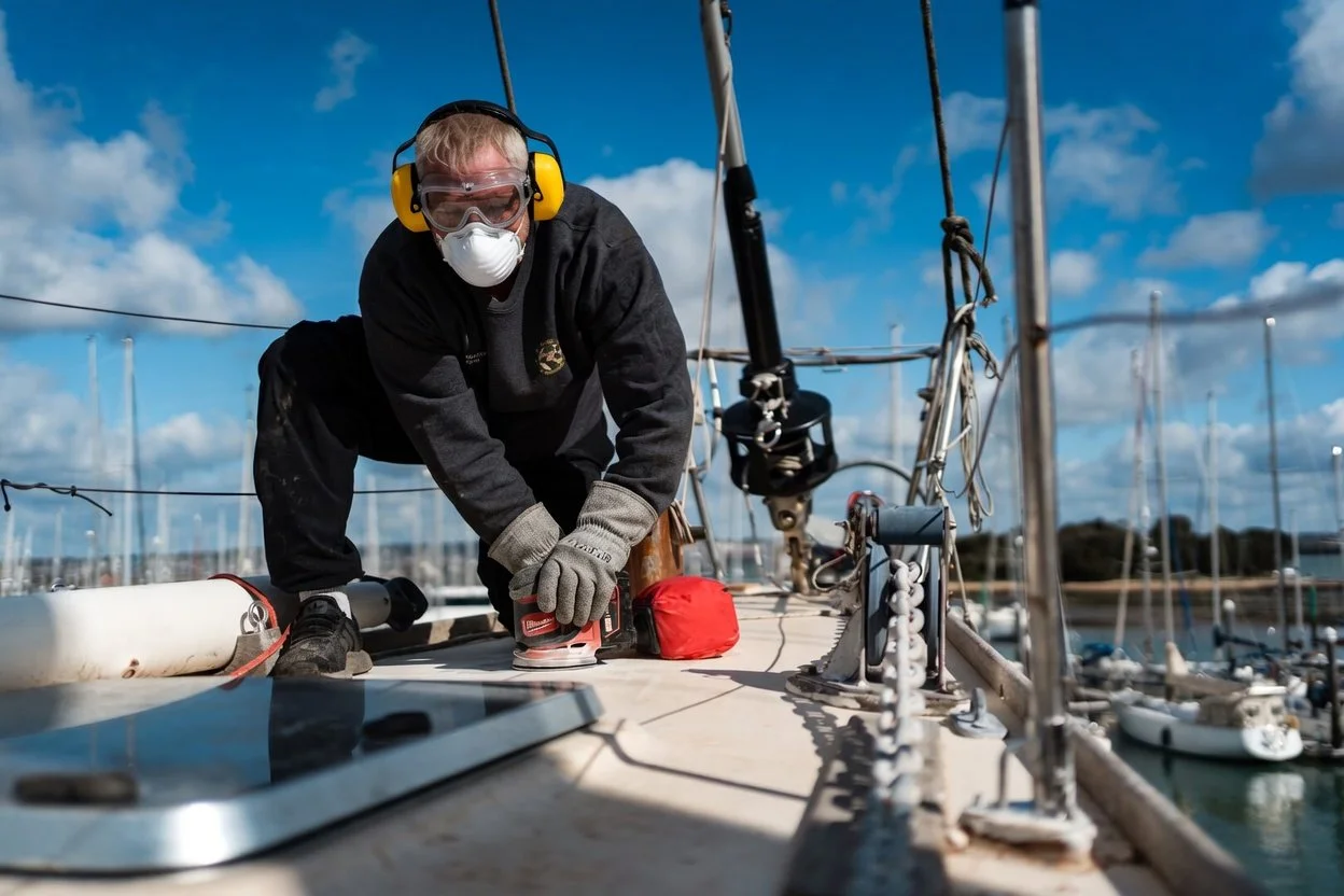A person working on a sailboat deck, wearing safety headphones, goggles, a mask, gloves, and dark clothing, with a marina filled with boats and blue sky with clouds in the background.