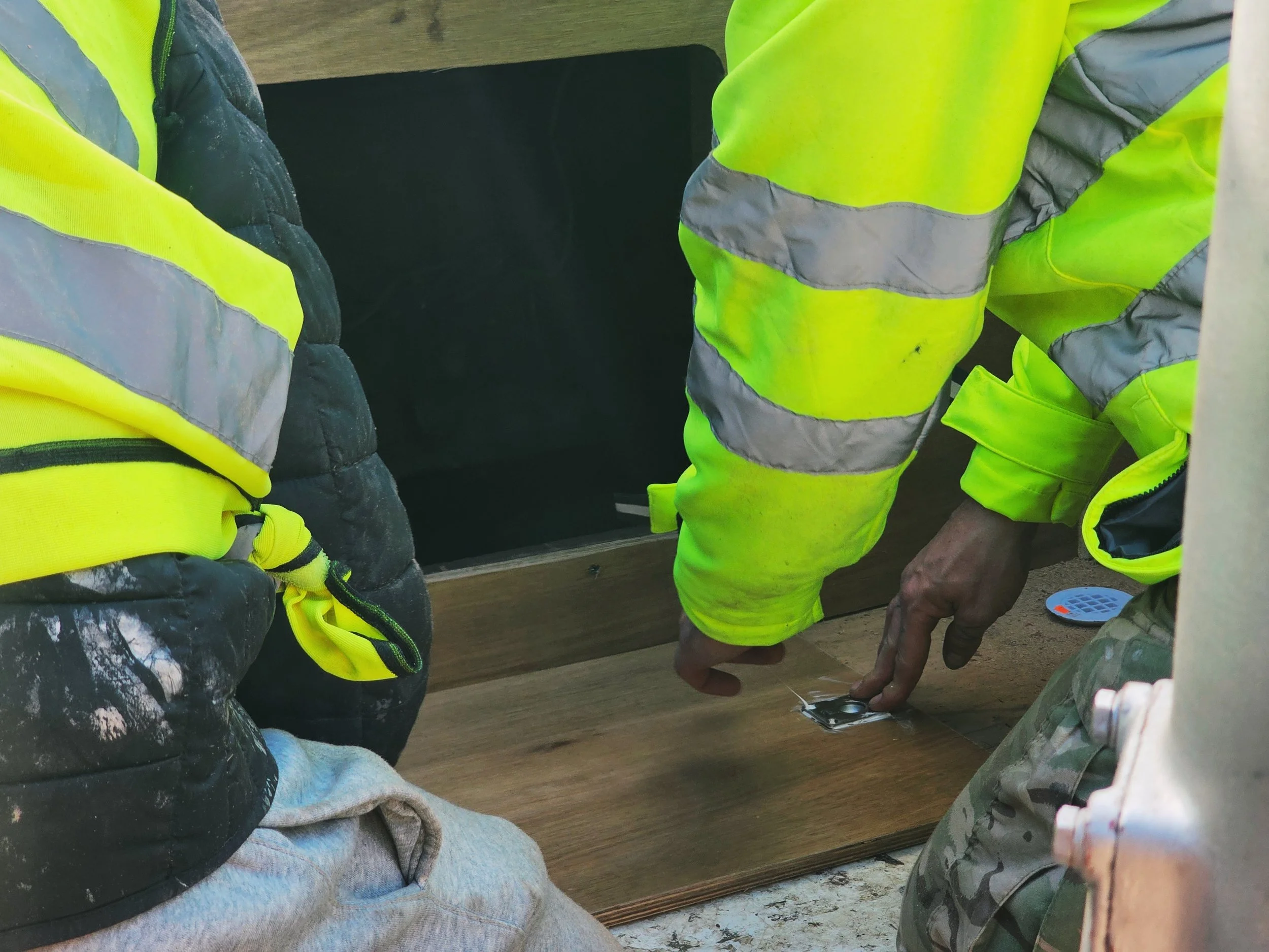 Two construction workers in high-visibility safety vests working on a wooden surface.