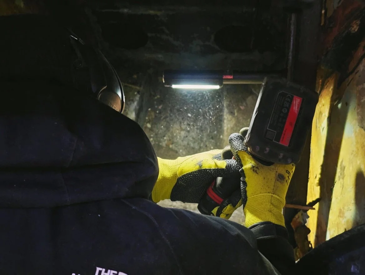 A worker in yellow gloves holding a digital device, working in a confined, dark space with a light overhead, possibly doing maintenance or inspection.