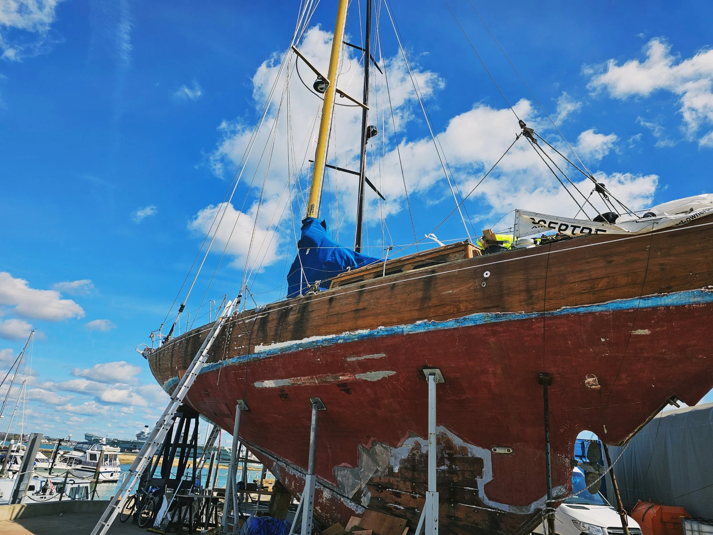 A wooden sailboat on dry dock with a climbing ladder leaning against it, surrounded by other boats in a marina, with a bright blue sky and scattered clouds overhead.
