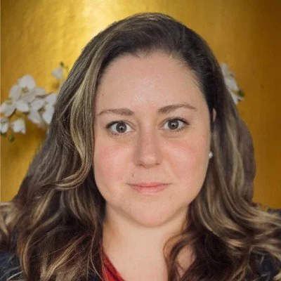 Close-up portrait of a woman with long wavy brown hair, wearing a red top, with a yellow background and white flowers.