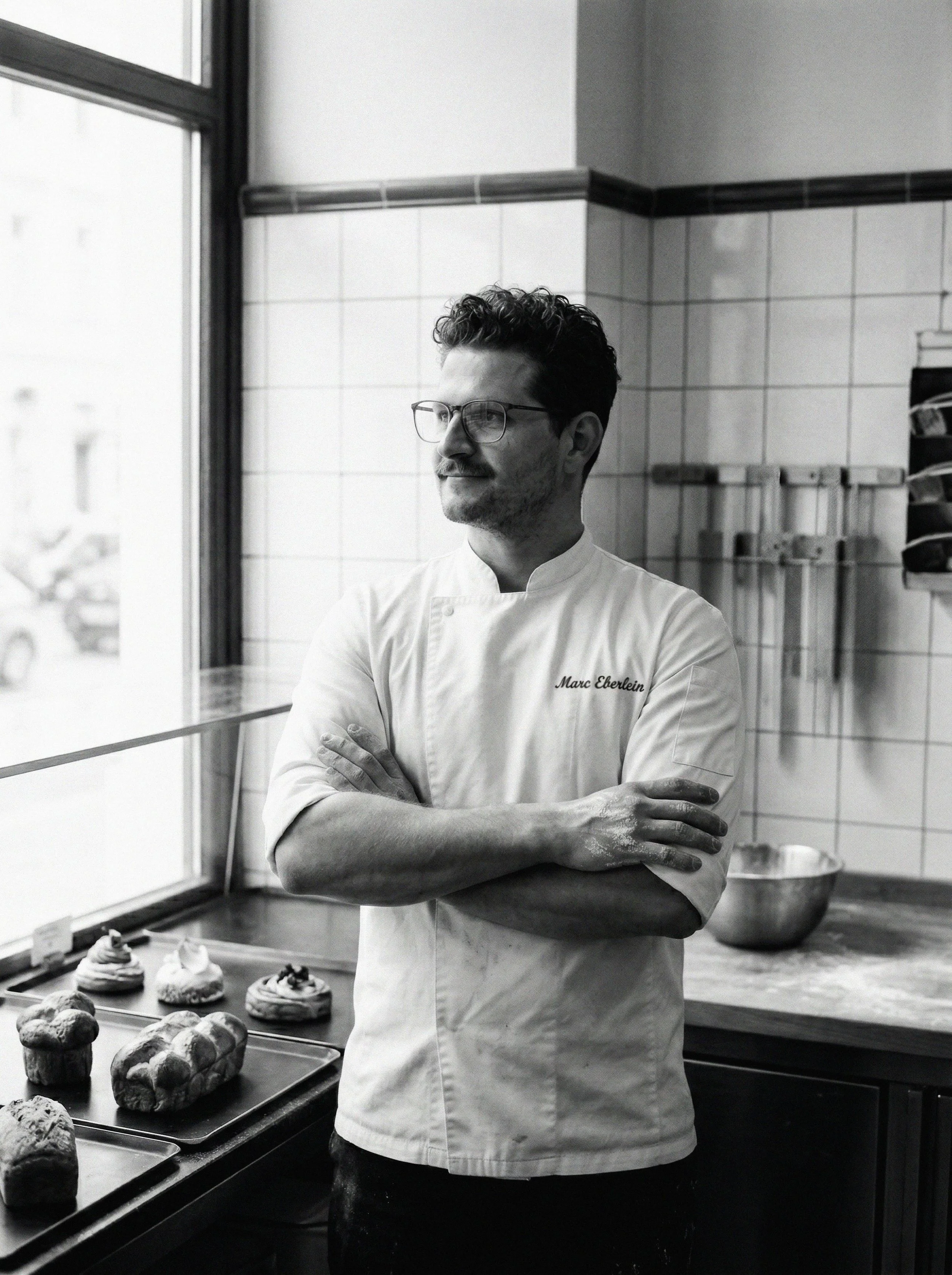 A male chef stands with arms crossed in a kitchen, wearing glasses and a white chef's coat with the name 'Marc Eberlein'. There are baked goods on a tray and a window behind him.